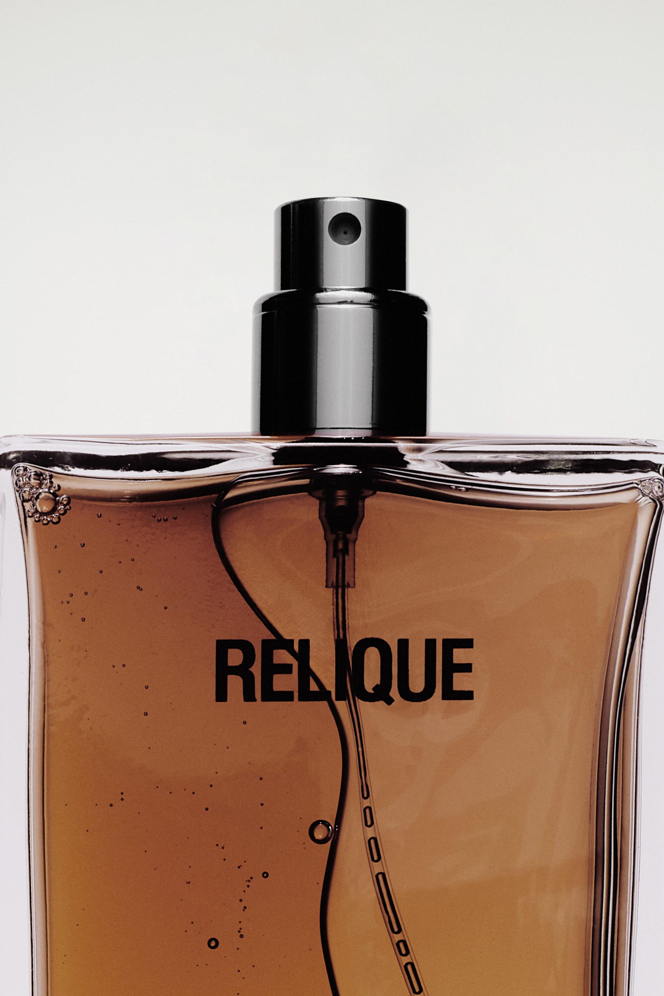 The COS Perfumery Eau de Parfum bottle is facing forwards, showing a close-up of its rectangular clear glass body filled with amber-coloured liquid. The bottle displays the bold black text 'RELIQUE' on the front, featuring a metallic black spray nozzle and a visible internal dip tube. The COS Perfumery Eau de Parfum bottle is facing forwards, showing a close-up of its rectangular clear glass body filled with amber-coloured liquid. The bottle displays the bold black text 'RELIQUE' on the front, featuring a metallic black spray nozzle and a visible internal dip tube.