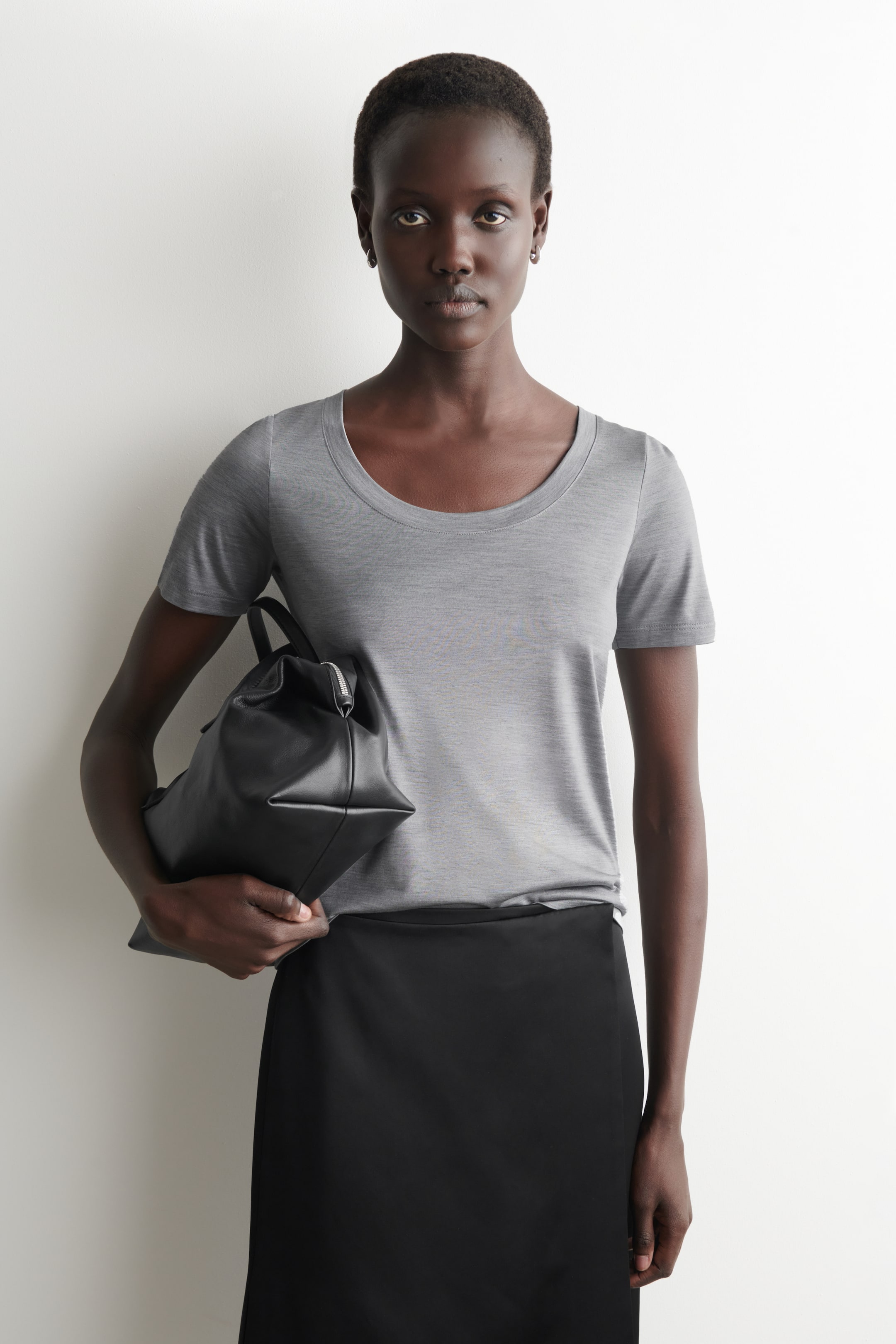 Agrandir l'image: Un t-shirt gris à manches courtes avec un col rond est porté, rentré dans une jupe noire taille haute présentant une coupe lisse et droite. Un sac structuré noir avec une fermeture éclair supérieure est tenu sous le bras.