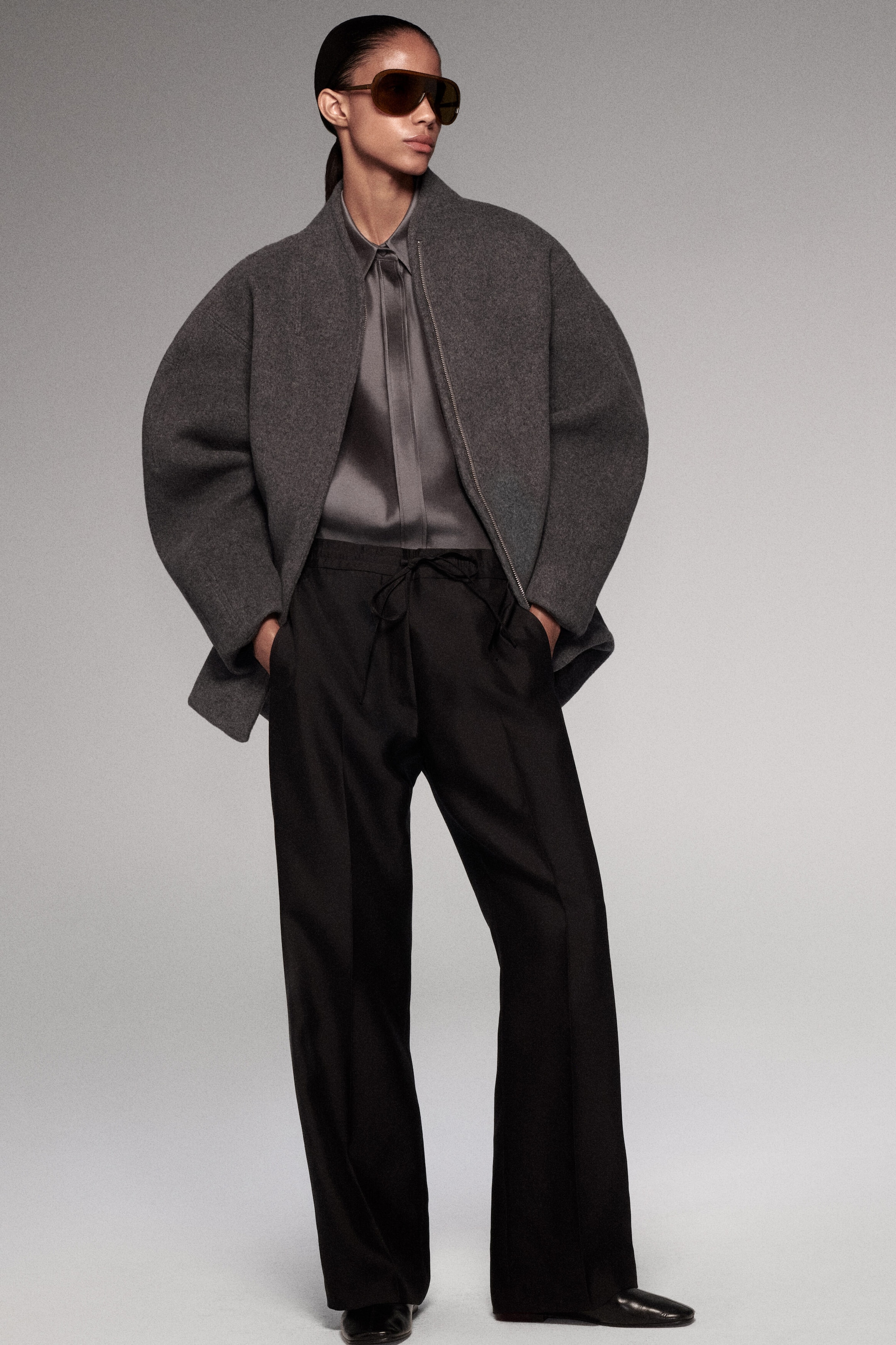 La veste zippée en mélange de laine à la coupe sculptée présente, de face, une silhouette surdimensionnée évoquant un cocon, avec des manches volumineuses et arrondies ainsi qu’un col montant. La veste grise texturée est superposée à une chemise en satin gris, mettant en valeur son drapé structuré et son tissu épais.