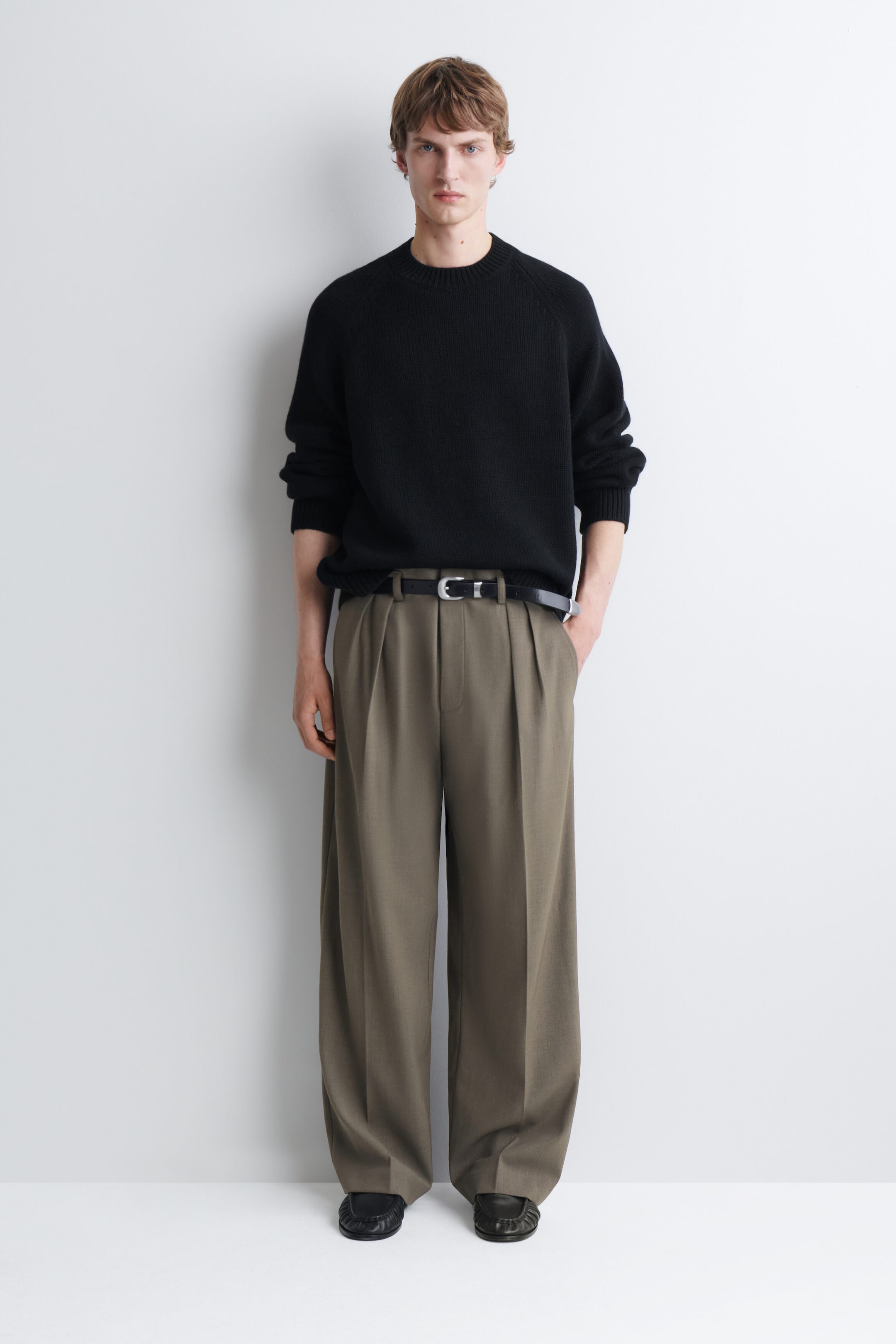Wide-leg pleated trousers in an olive-brown colour are being worn by the model, styled with a black leather belt and a black crew-neck jumper. The trousers feature deep front pleats and a relaxed fit that drapes down to the shoes. Wide-leg pleated trousers in an olive-brown colour are being worn by the model, styled with a black leather belt and a black crew-neck jumper. The trousers feature deep front pleats and a relaxed fit that drapes down to the shoes.