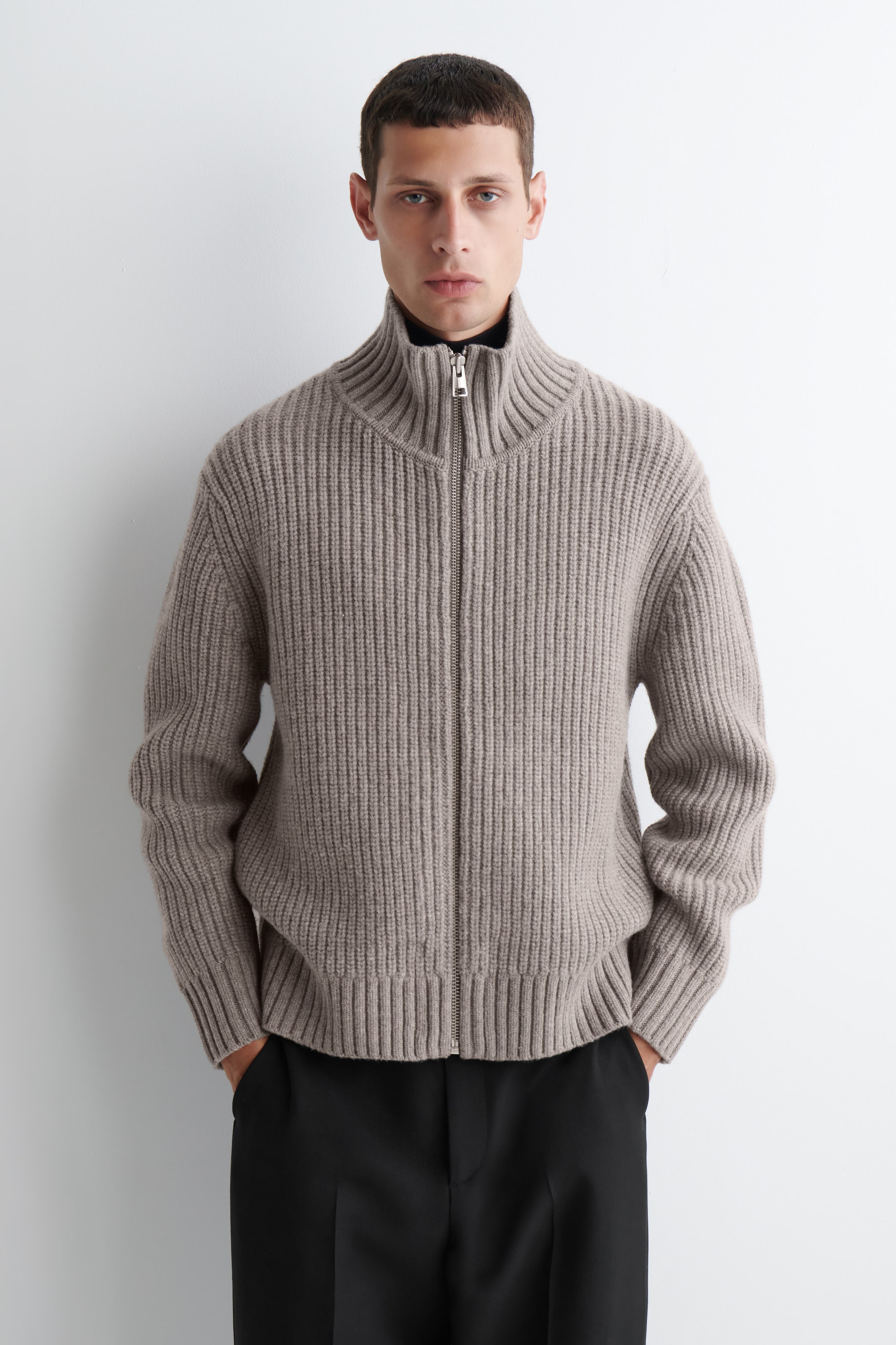 Se større billede: En taupe ribbet sweater med fuld lynlås vender mod fronten, med en stående høj krave og en sølvfarvet lynlås, der løber ned langs fronten. Fremtrædende lodrette ribber er synlige over hele sweateren, med en sort højnekket top, der kommer til syne under dens delvist åbnede krave.