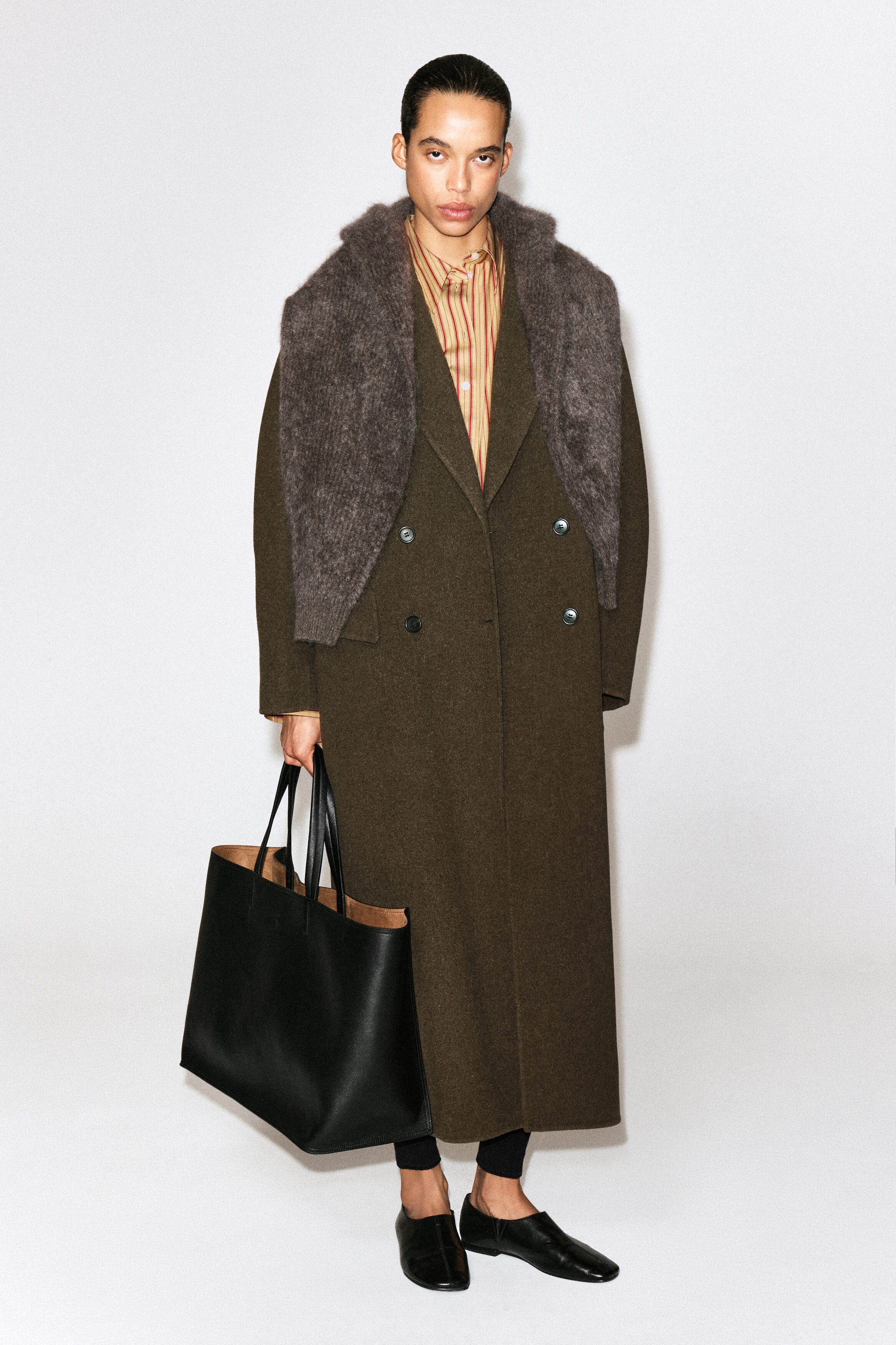 The striped cotton-lyocell shirt, featuring vertical red and yellow-beige stripes, is worn from the front, layered beneath a double-breasted coat and a draped knit scarf. Only the classic collar and upper buttoned placket of the shirt are visible, showcasing its structured design.