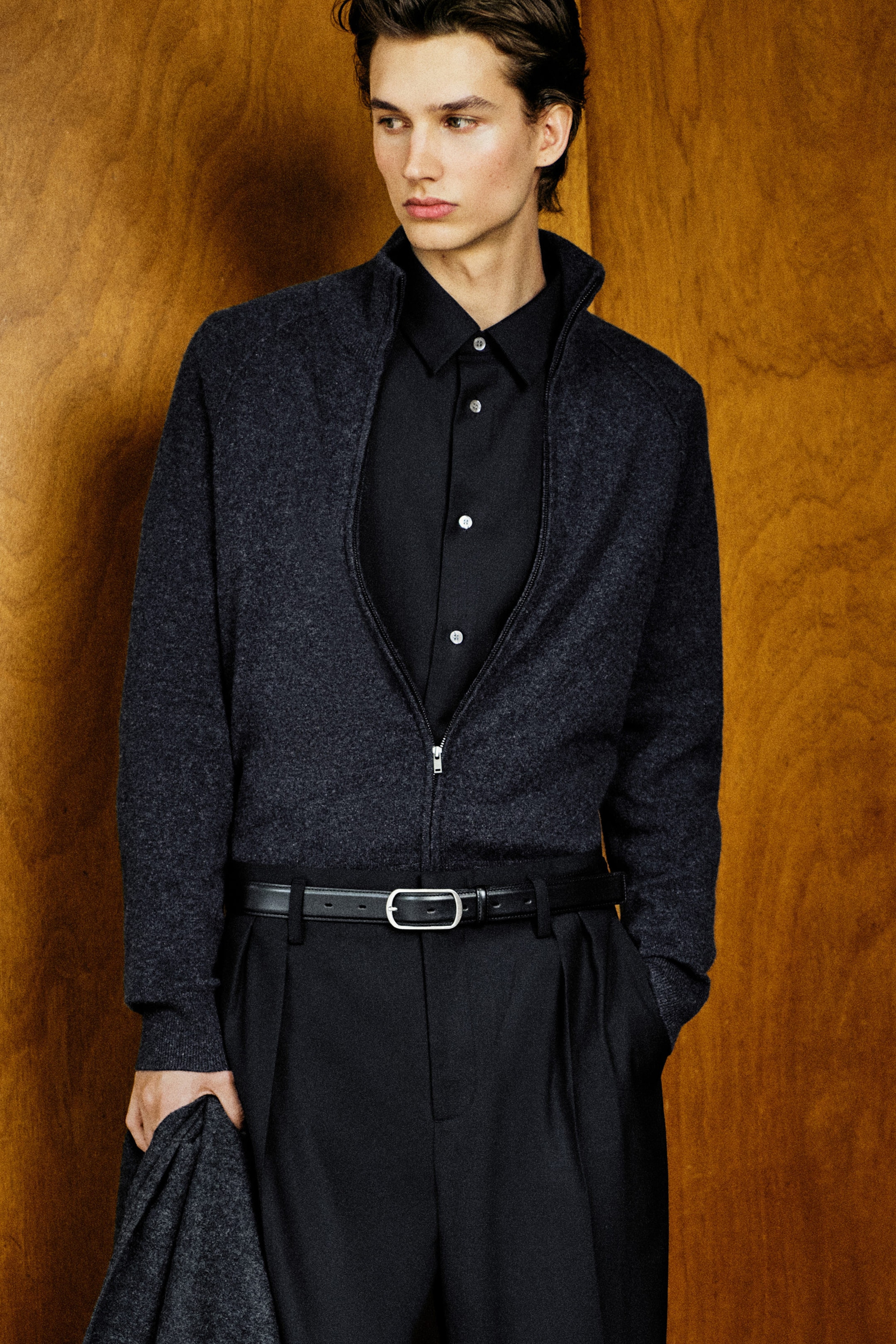 The dark grey cashmere funnel-neck zip-up cardigan is shown from the front, featuring a high collar and a silver-tone zip. The cardigan is worn partially zipped over a black button-up shirt and tucked into black trousers, highlighting its soft knit texture and ribbed cuffs. The dark grey cashmere funnel-neck zip-up cardigan is shown from the front, featuring a high collar and a silver-tone zip. The cardigan is worn partially zipped over a black button-up shirt and tucked into black trousers, highlighting its soft knit texture and ribbed cuffs.