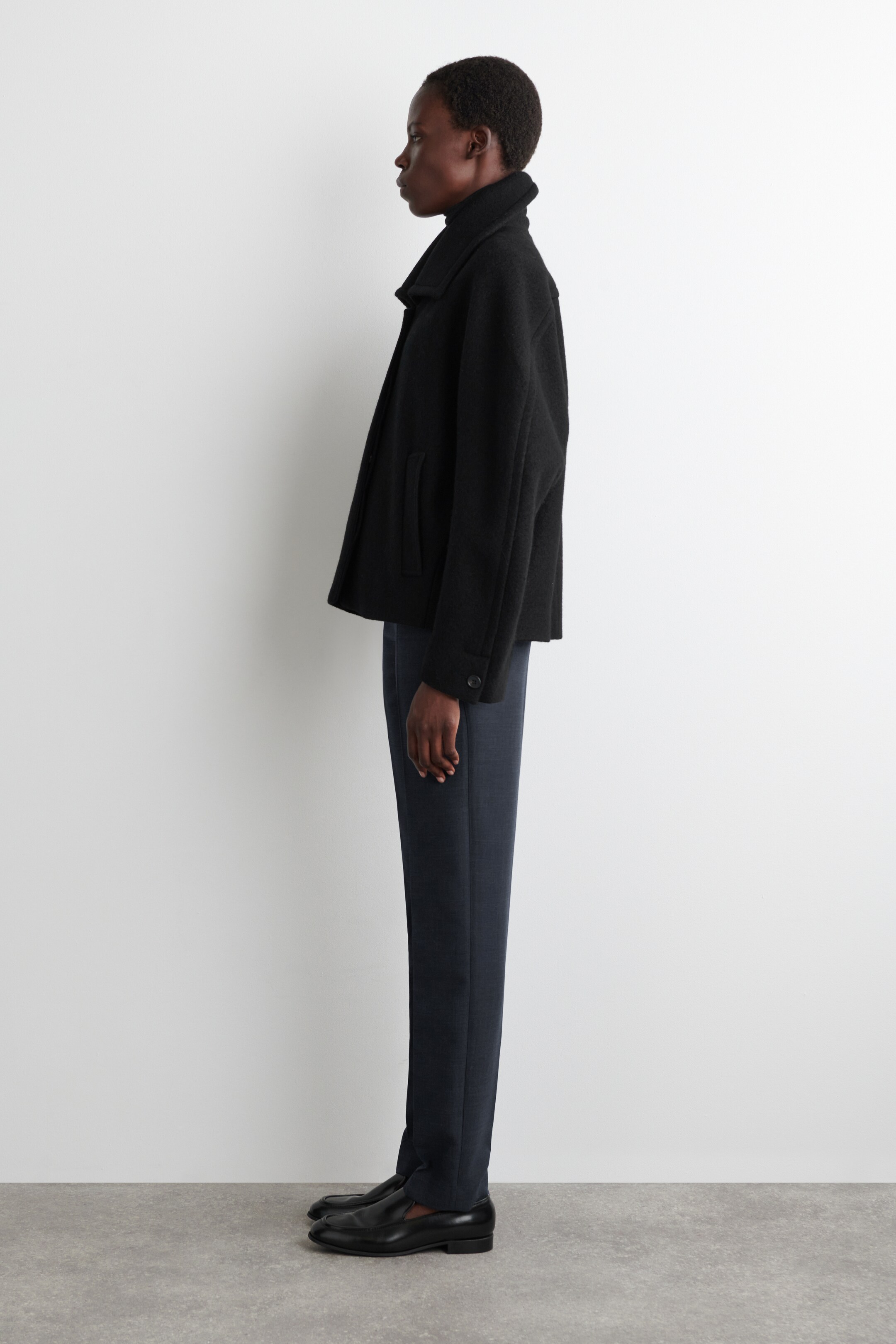 Grotere afbeelding bekijken: Een zwarte korte jas wordt getoond vanuit zijn zijprofiel, met een kraag in peacoat-stijl, zichtbare paspelzakken en knoopmanchetten. Het wordt gestyled met donkerblauwe broek en zwarte loafers.
