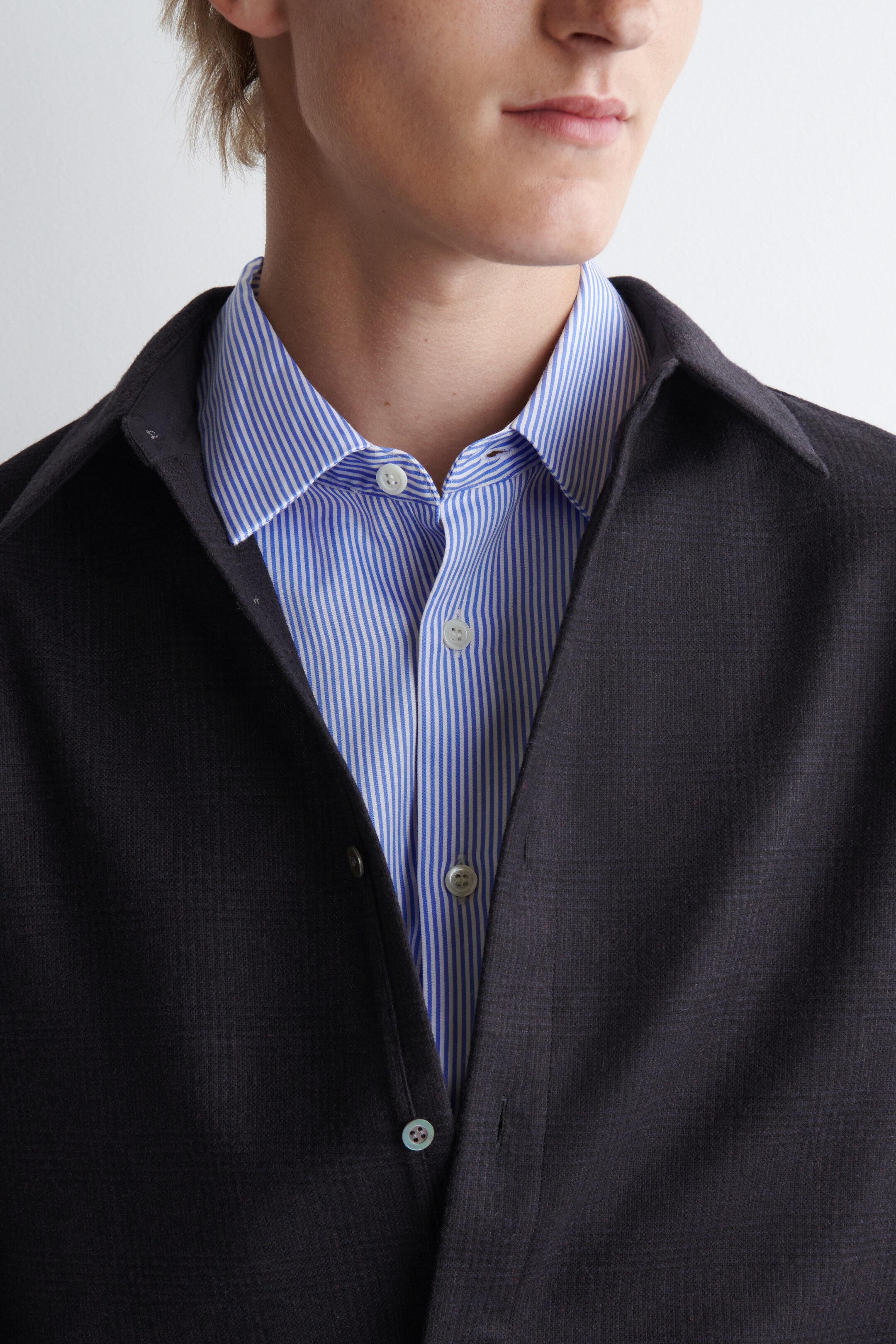 View larger image: A dark gray jacket, displaying a subtle check pattern, is layering over a blue and white vertically striped button-up shirt. The jacket is featuring an open front with visible light-colored buttons, and the shirt is showing a classic collar with its top buttons unfastened.