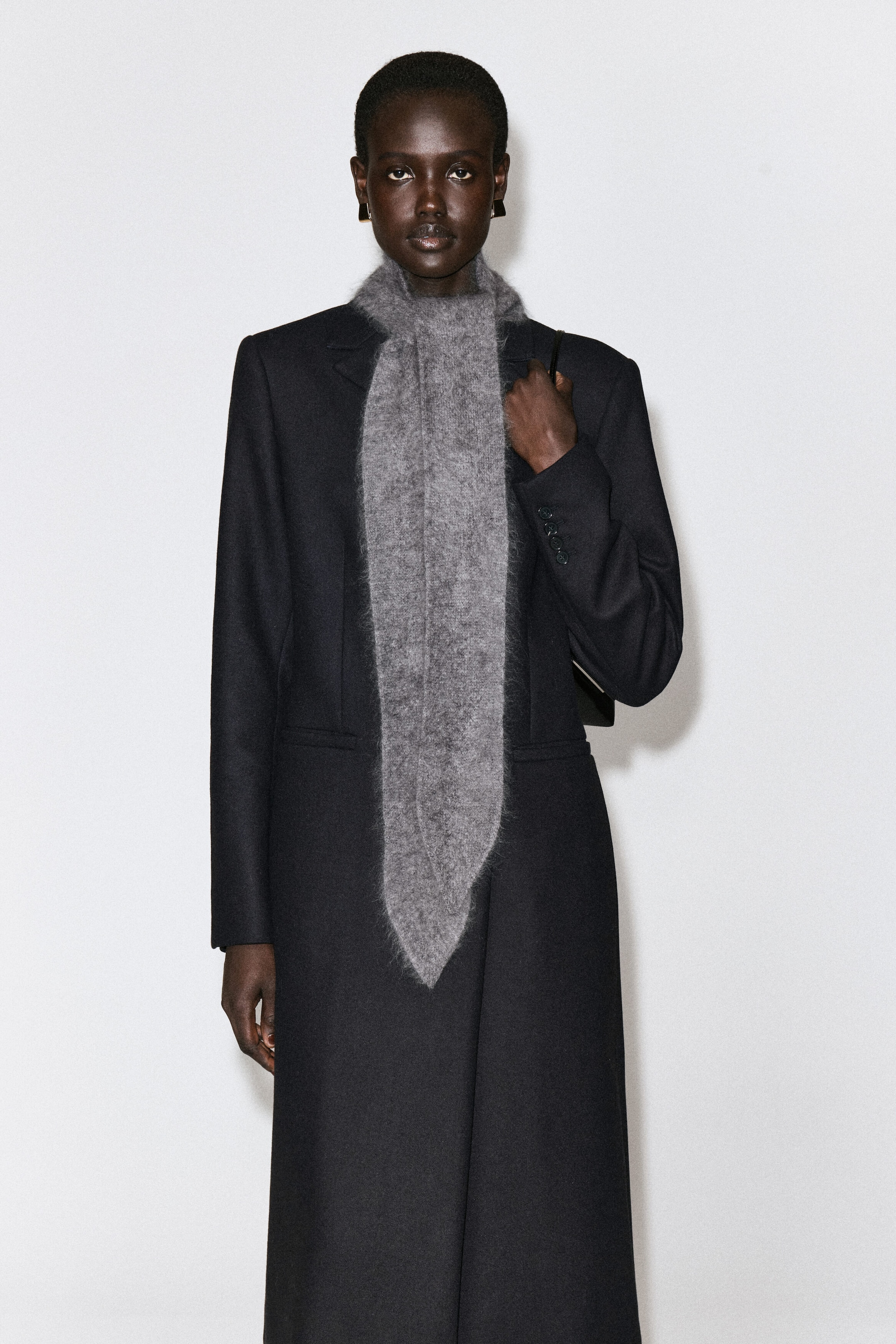 A gray brushed-wool scarf is shown from the front, draped over a tailored black coat and tied like a classic necktie. The scarf features a fuzzy, textured knit with tapered, pointed ends hanging down the front. A gray brushed-wool scarf is shown from the front, draped over a tailored black coat and tied like a classic necktie. The scarf features a fuzzy, textured knit with tapered, pointed ends hanging down the front.