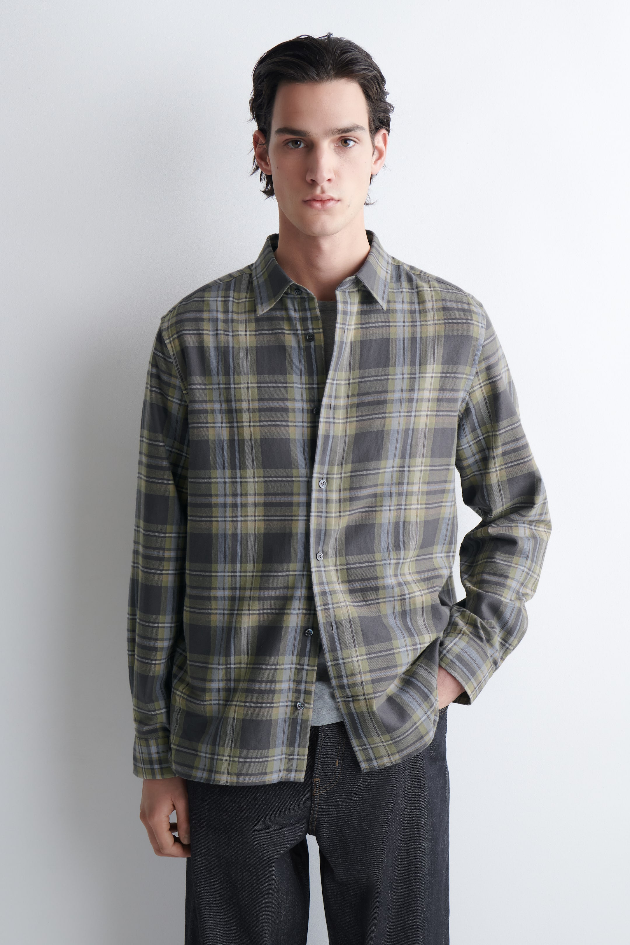 Agrandir l'image: Une chemise à carreaux à manches longues est présentée de face, arborant un motif à carreaux dans des tons de gris, vert olive et bleu clair. Elle est conçue avec un col pointu, une fermeture boutonnée à l'avant, et des manches longues avec des poignets boutonnés, affichant une coupe régulière et portée ouverte sur un sous-vêtement gris.