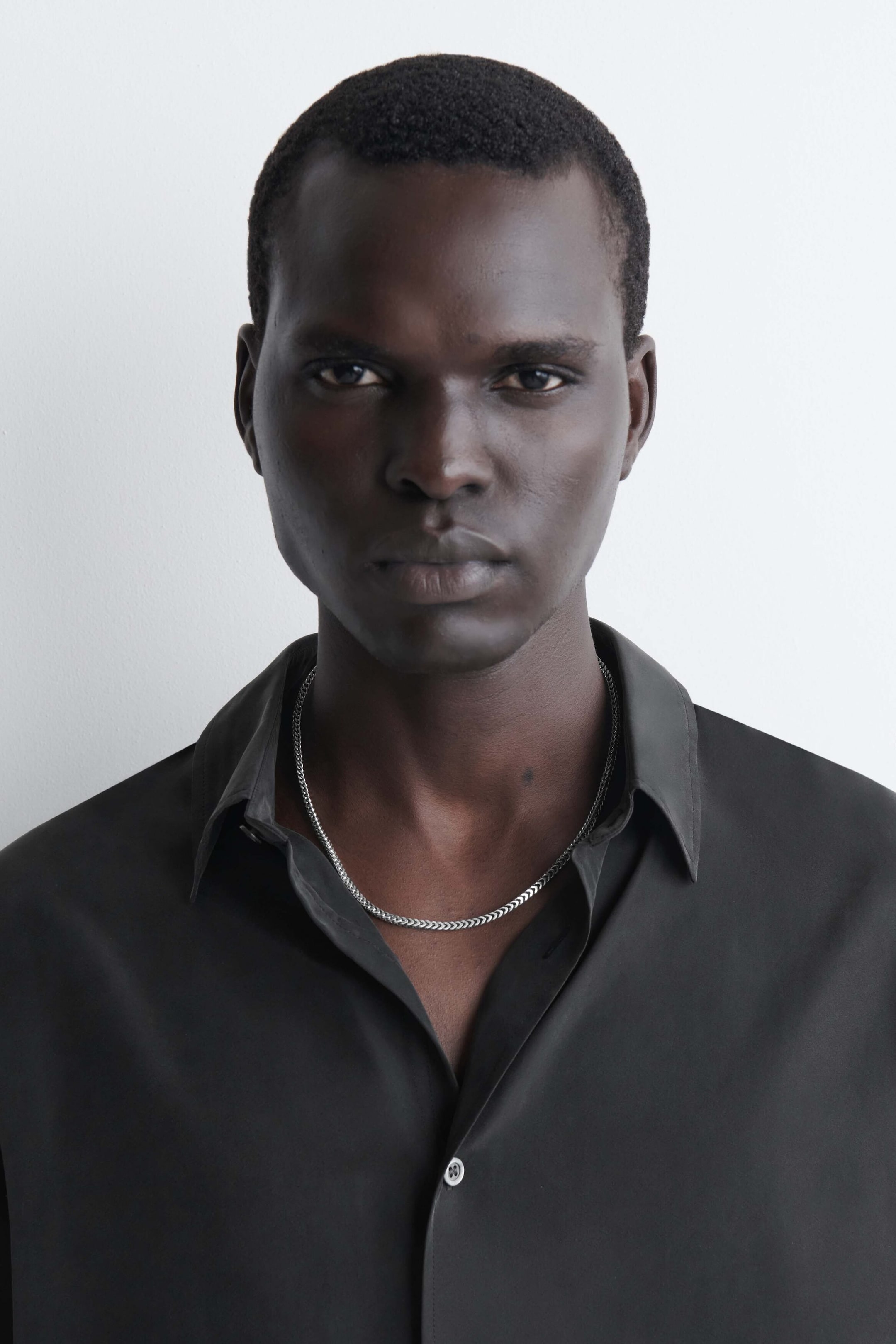 A silver-toned chain necklace featuring a textured, chevron-patterned link design rests around the neck, facing forward. The necklace is styled under the open collar of a dark grey button-up shirt, highlighting its metallic finish.