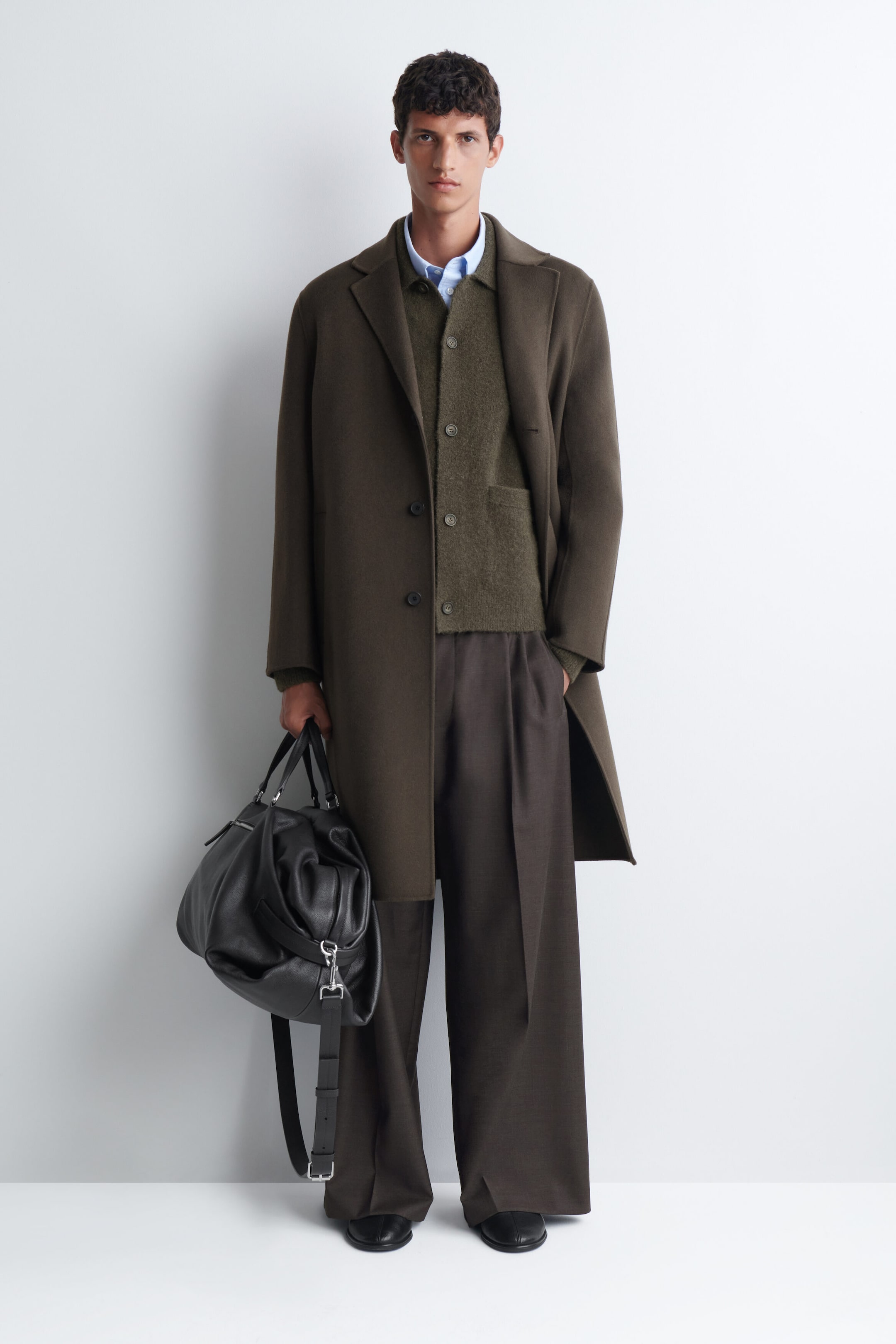 The model is wearing a textured, collared cardigan in olive green, layered over a light blue collared shirt and under a dark olive-brown overcoat. The cardigan features a button-front closure, a flat collar, and a soft, fuzzy knit texture. The model is wearing a textured, collared cardigan in olive green, layered over a light blue collared shirt and under a dark olive-brown overcoat. The cardigan features a button-front closure, a flat collar, and a soft, fuzzy knit texture.