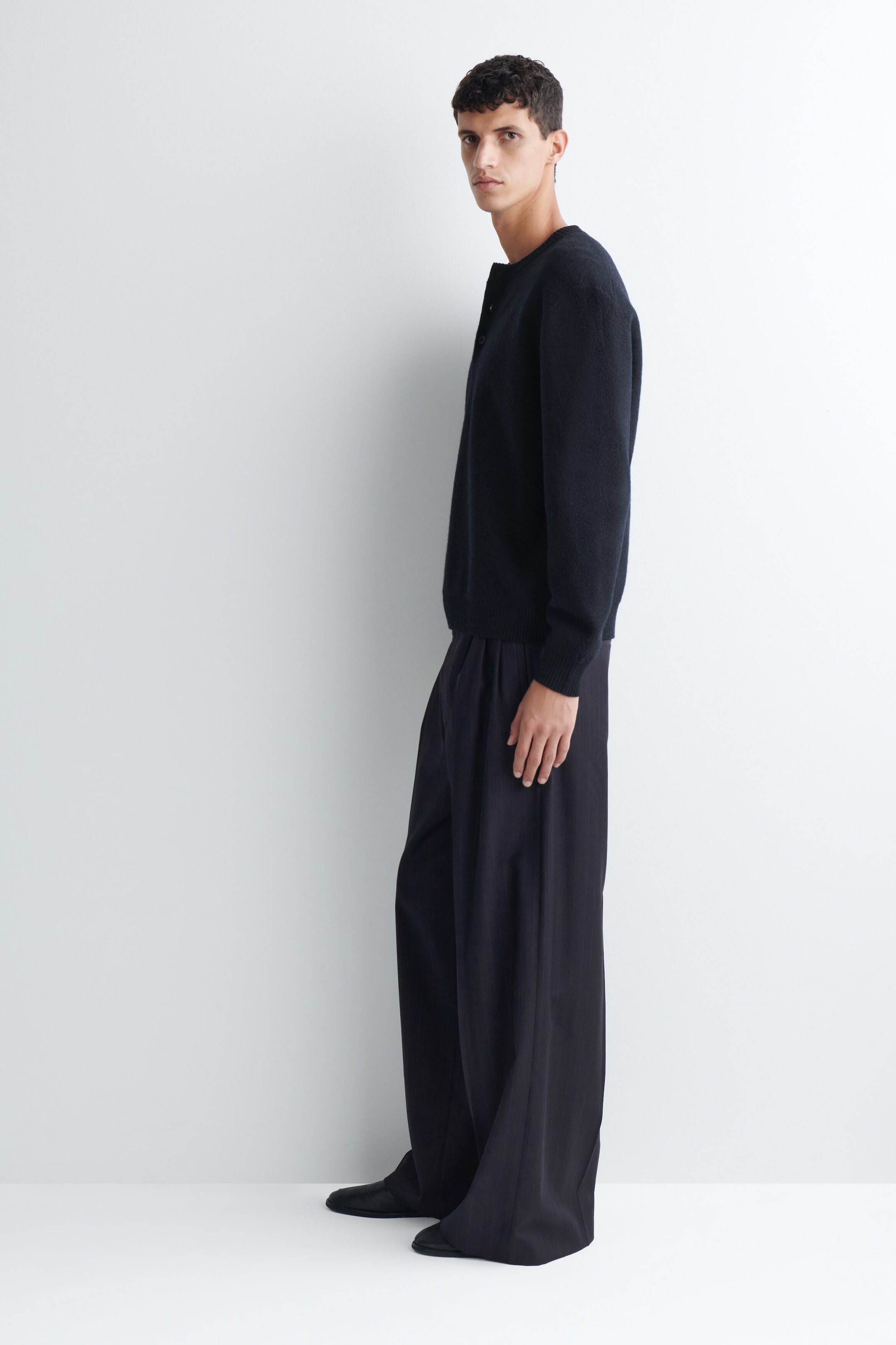 Pantalon plissé à jambe large et coupe décontractée, en gris foncé, porté par le mannequin, mettant en valeur une silhouette drapée et de profondes plis à l’avant. Le pantalon est agencé avec un haut en tricot à manches longues foncé et des chaussures en cuir noires. Pantalon plissé à jambe large et coupe décontractée, en gris foncé, porté par le mannequin, mettant en valeur une silhouette drapée et de profondes plis à l’avant. Le pantalon est agencé avec un haut en tricot à manches longues foncé et des chaussures en cuir noires.