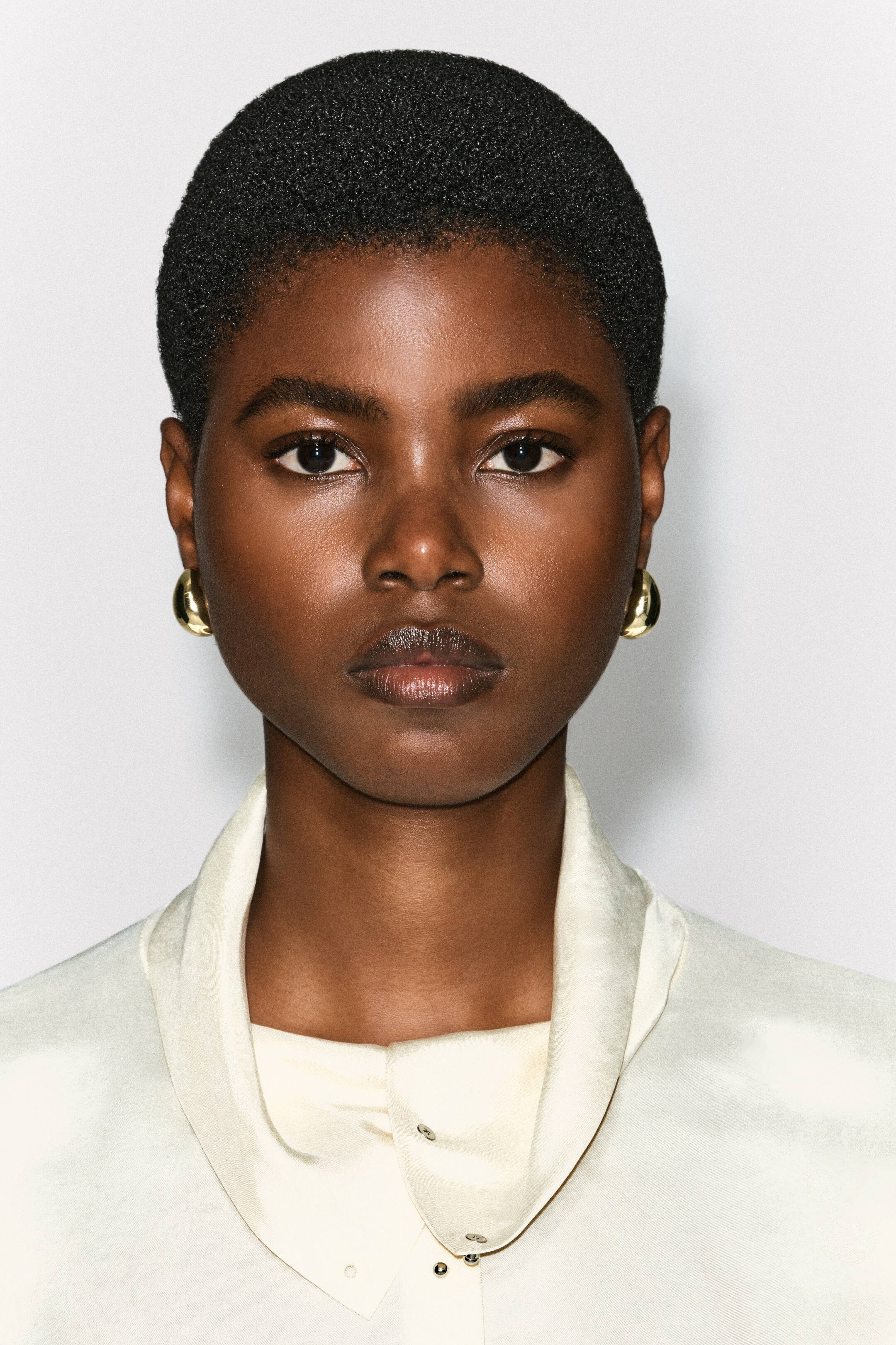Agrandir l'image: Des boucles d’oreilles bombées incurvées à finition dorée polie sont portées par le mannequin, affichant un design lisse et arrondi qui épouse le lobe de l’oreille.