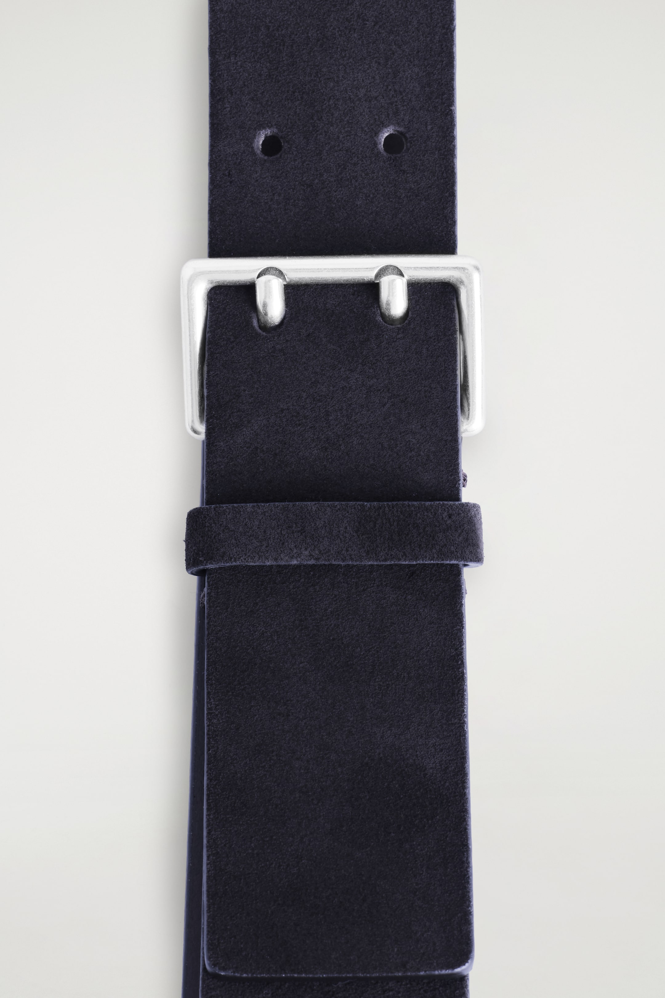 View larger image: A dark blue suede belt is being shown from a top view, featuring a squared, silver-tone double-prong buckle with two visible belt holes above it, and a matching suede keeper loop securing the strap below.