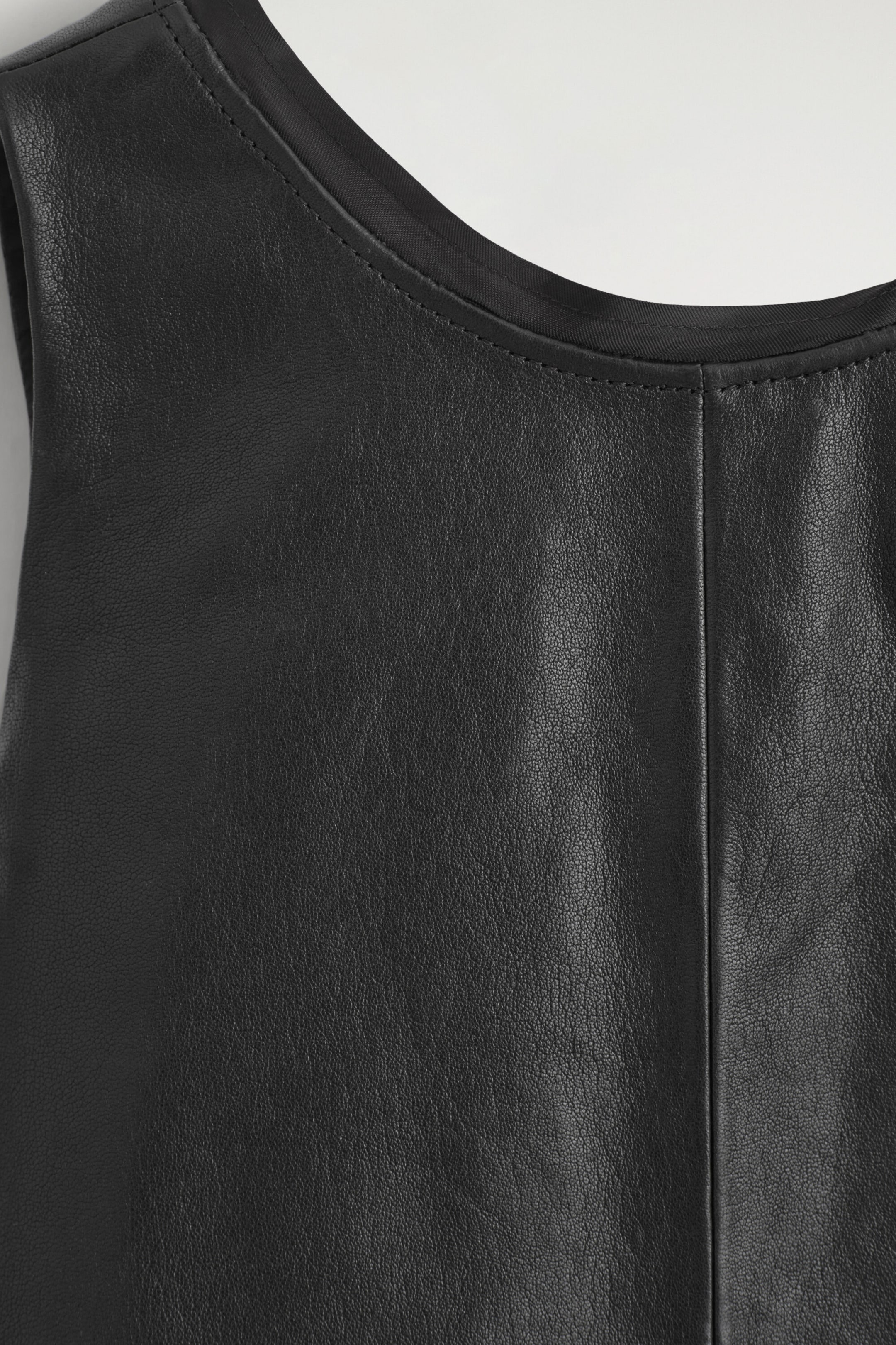 Ein schwarzes Leder-Mini-A-Linienkleid wird in einer Nahaufnahme von vorne gezeigt und präsentiert die feinkörnige Textur des Leders sowie eine markante vertikale Mittelnaht. Das Kleid hat einen U-Ausschnitt mit sichtbaren Steppnähten und einer mit Stoff eingefassten Innenkante. Ein schwarzes Leder-Mini-A-Linienkleid wird in einer Nahaufnahme von vorne gezeigt und präsentiert die feinkörnige Textur des Leders sowie eine markante vertikale Mittelnaht. Das Kleid hat einen U-Ausschnitt mit sichtbaren Steppnähten und einer mit Stoff eingefassten Innenkante.
