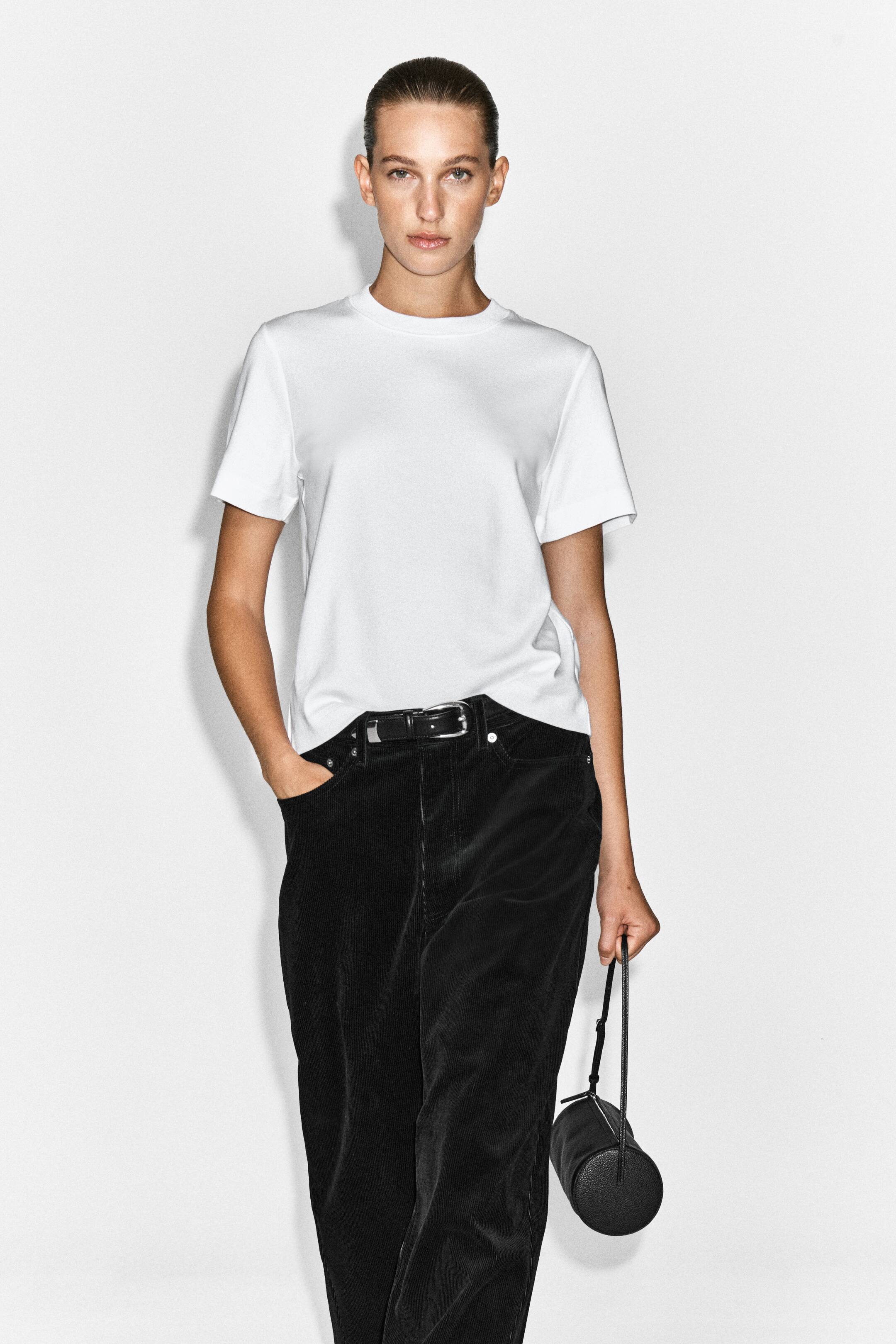 Visualizza l'immagine ingrandita: La modella indossa una t-shirt bianca con girocollo, infilata in pantaloni di velluto a coste neri a vita alta con cintura con fibbia argentata. La modella tiene in una mano una piccola borsa cilindrica nera, mentre appoggia l'altra mano nella tasca dei pantaloni.