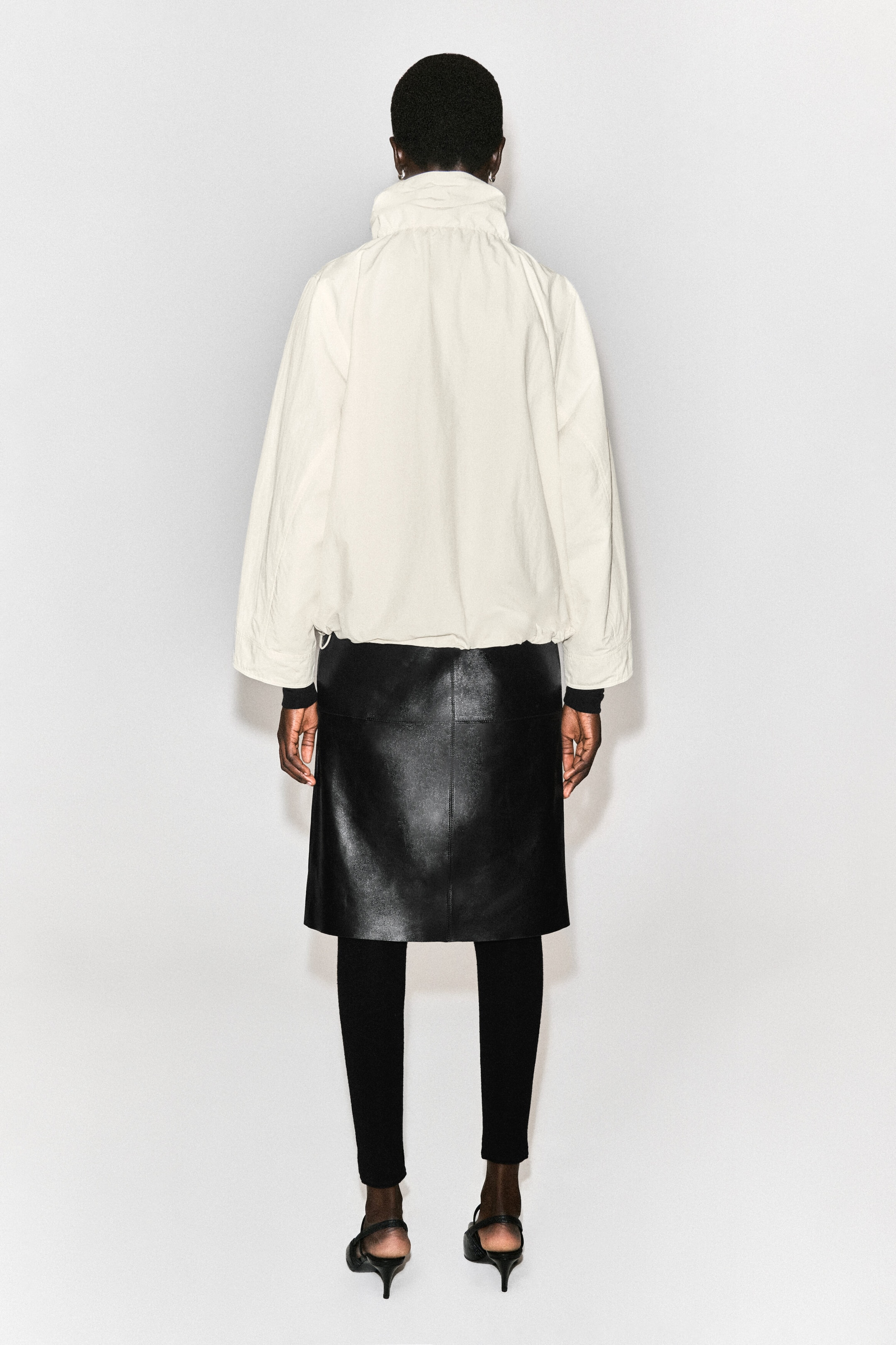 La veste en nylon à col cheminée est montrée de dos, mettant en valeur sa silhouette décontractée blanc cassé avec épaules tombantes et manches larges. Elle présente un col cheminée haut froncé et un ourlet légèrement froncé et élastiqué. La veste en nylon à col cheminée est montrée de dos, mettant en valeur sa silhouette décontractée blanc cassé avec épaules tombantes et manches larges. Elle présente un col cheminée haut froncé et un ourlet légèrement froncé et élastiqué.