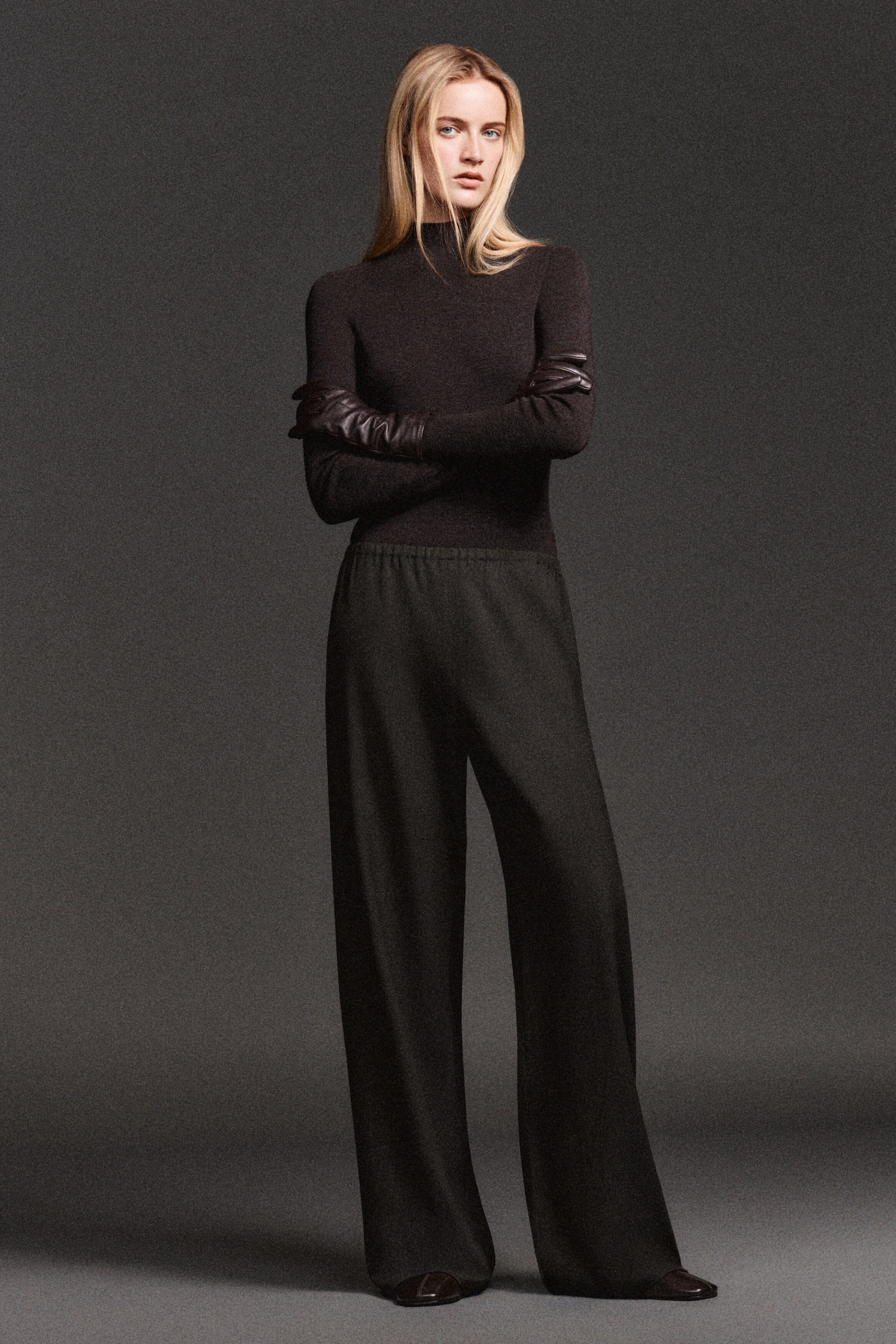 Facing the front, the model is wearing a dark brown monochromatic ensemble featuring a fitted mock-neck knit top and high-waisted, wide-leg trousers. Long, dark brown leather gloves are styled over the forearms of the long-sleeved top, complementing the minimalist design. Facing the front, the model is wearing a dark brown monochromatic ensemble featuring a fitted mock-neck knit top and high-waisted, wide-leg trousers. Long, dark brown leather gloves are styled over the forearms of the long-sleeved top, complementing the minimalist design.