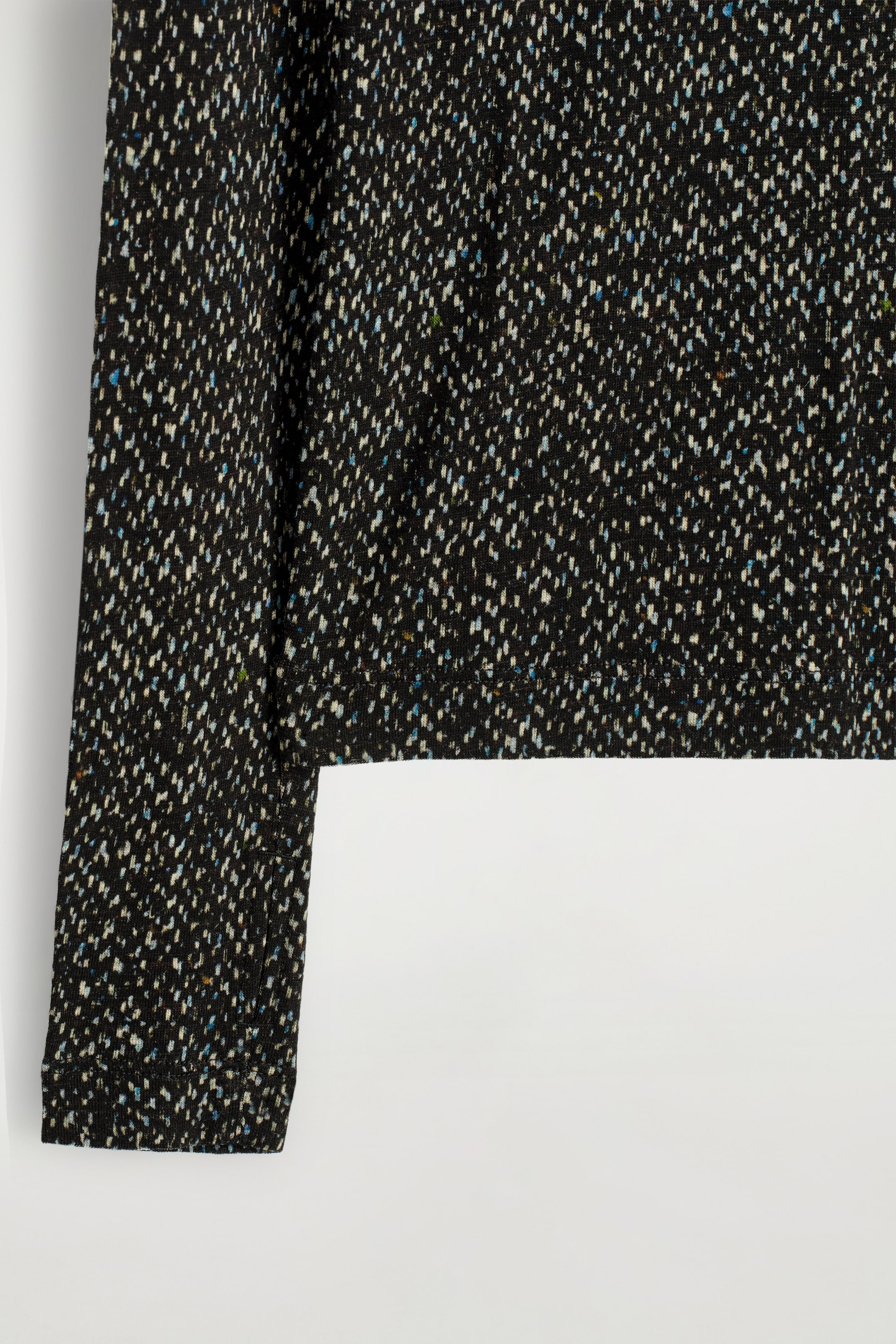 Grösseres Bild ansehen: Ein schwarzes langärmliges Oberteil zeigt eine fein gestrickte Textur mit einem gesprenkelten Muster aus weißen und hellblauen Sprenkeln.