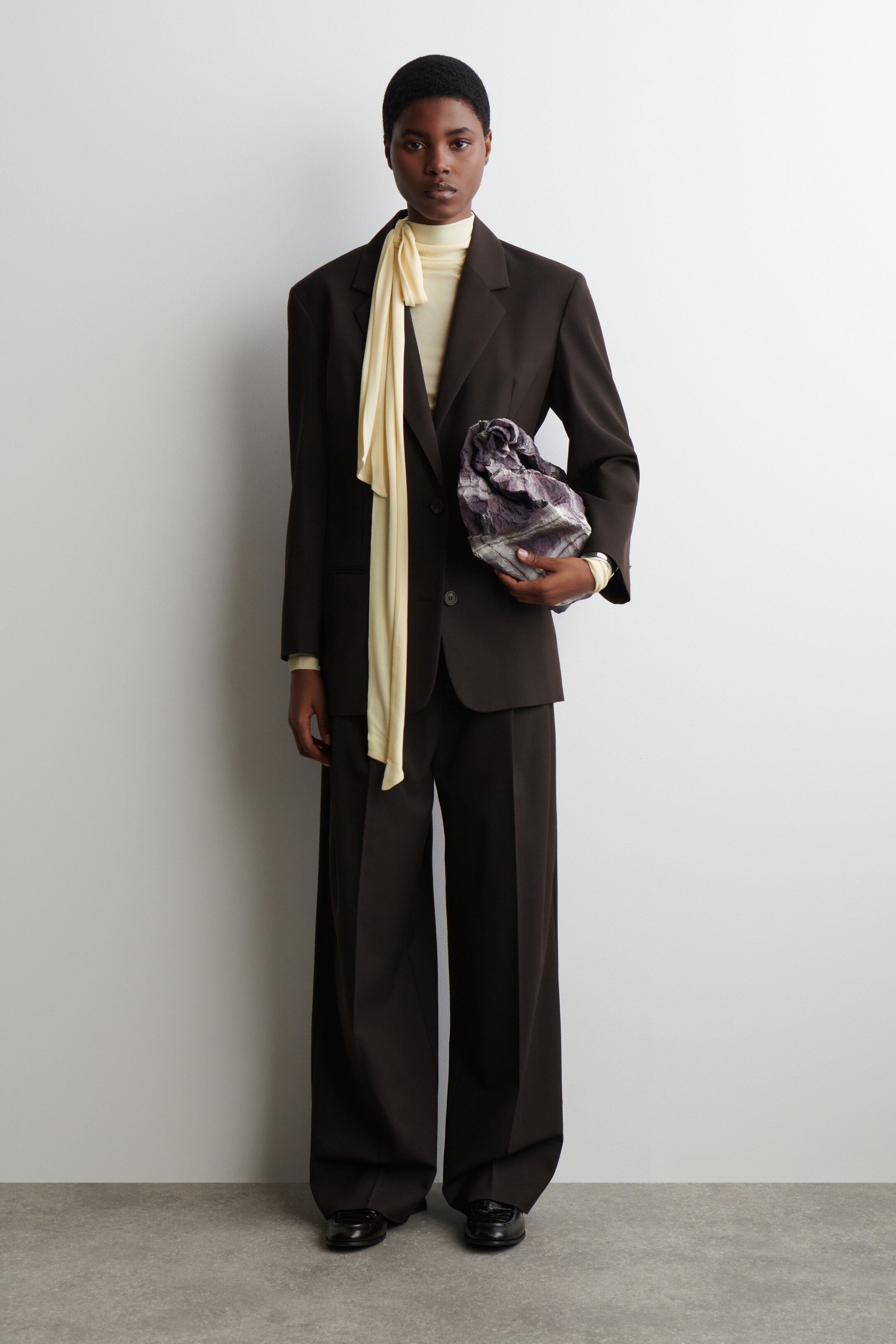 Grösseres Bild ansehen: Ein dunkelbrauner Anzug, bestehend aus einem einreihigen Blazer mit breiten Schultern und Kerbrevers sowie einer Hose mit weitem Bein, wird über einem hellgelben Rollkragenoberteil getragen, mit einem passenden langen Schal, der um den Hals drapiert ist. Eine geraffte, marmorierte lila-graue Tasche wird in einer Hand gehalten, und schwarze flache Schuhe sind an den Füßen sichtbar.