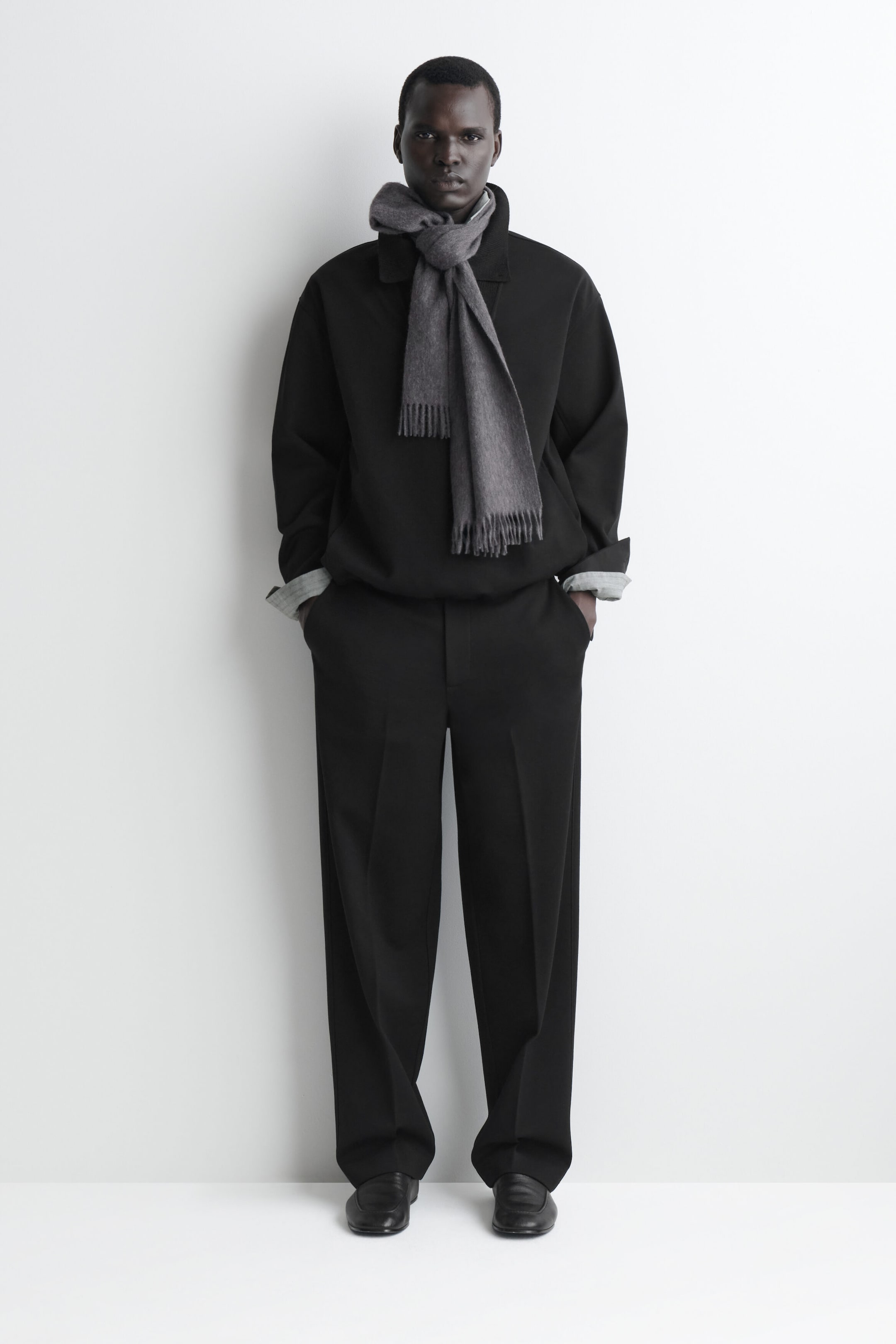 A grey fringed cashmere scarf is shown from the front, looped around the model’s neck and draped over a black pullover. The scarf features a soft woven texture and fine fringe detailing at both ends. A grey fringed cashmere scarf is shown from the front, looped around the model’s neck and draped over a black pullover. The scarf features a soft woven texture and fine fringe detailing at both ends.