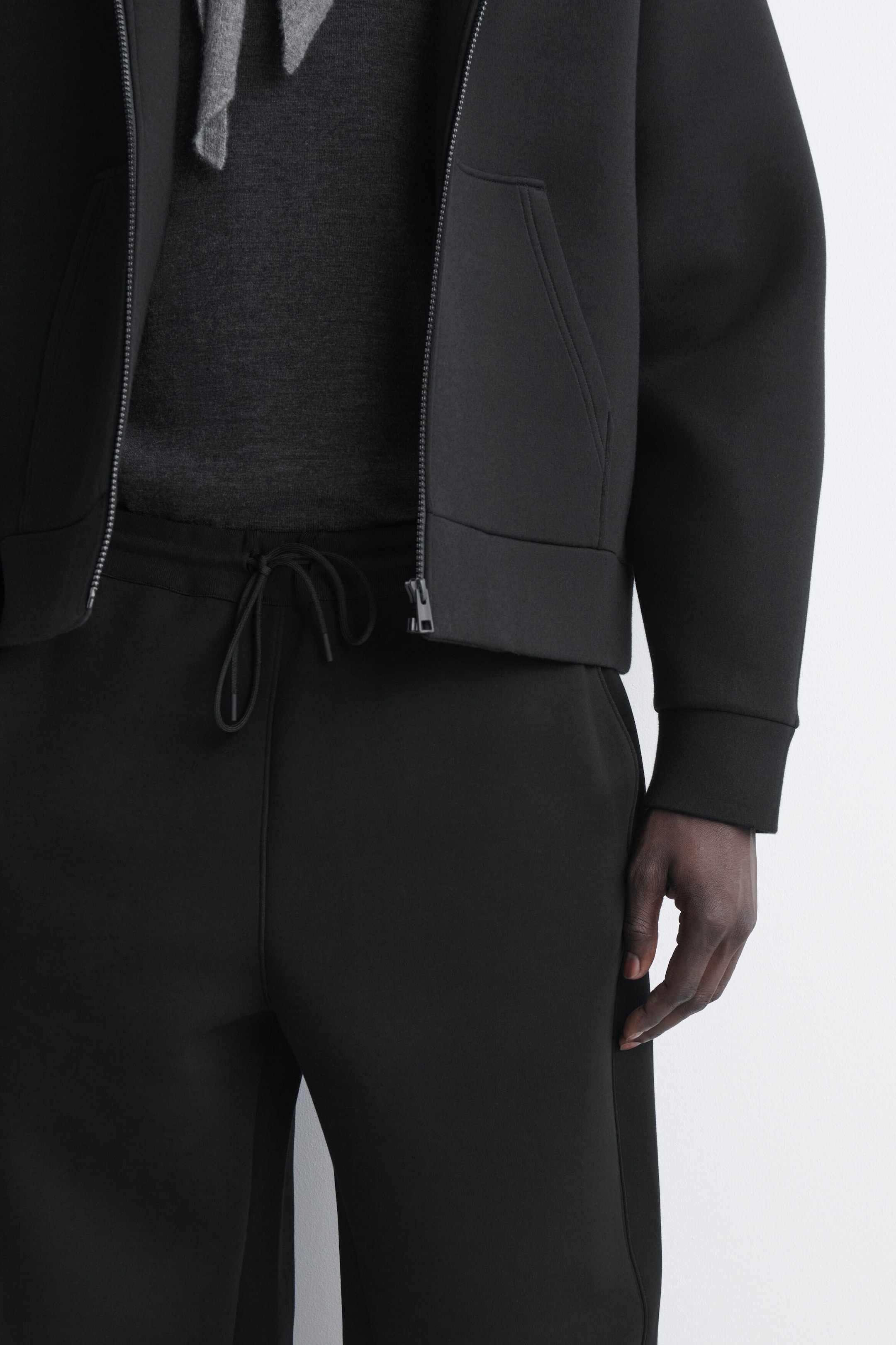 A black zip-up jacket and matching sweatpants are worn in a front-facing view. The jacket showcases a smooth, structured fabric with an open front zipper, ribbed cuffs, and side pockets, layered over a dark gray top. The matching sweatpants feature an elasticated waistband with an adjustable drawstring tie at the front.