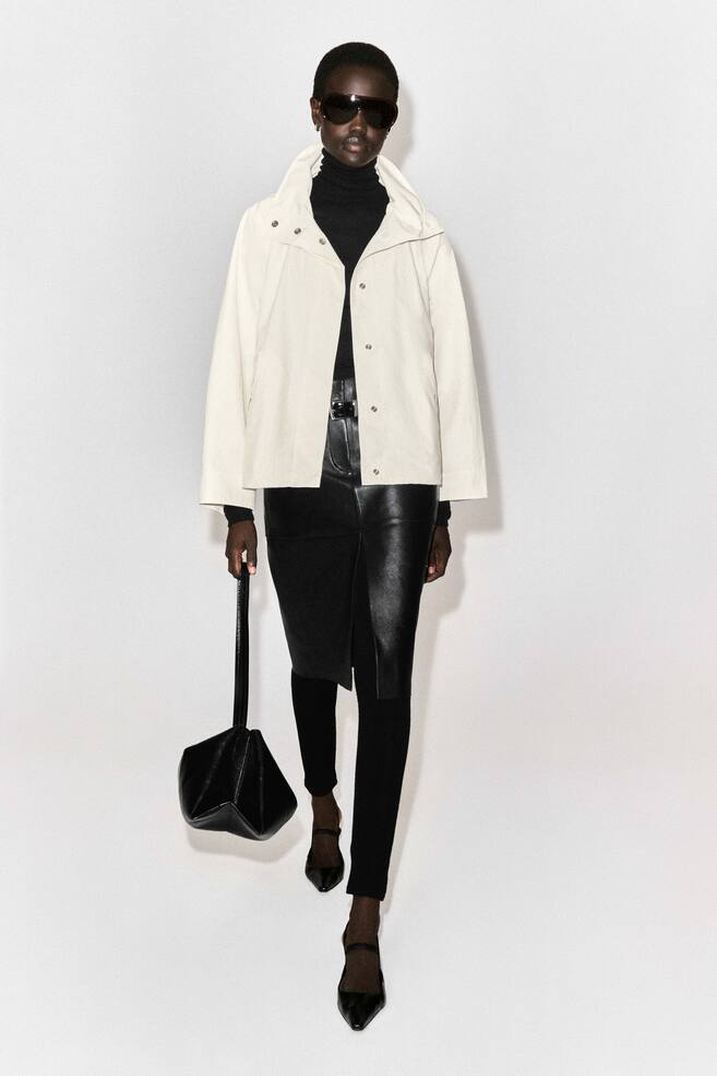 The off-white nylon funnel-neck jacket is shown from the front, featuring a relaxed fit, a high stand-up collar with snap fasteners, and a straight hem. The jacket is worn open over a black roll-neck, highlighting its clean, structured silhouette. - 1