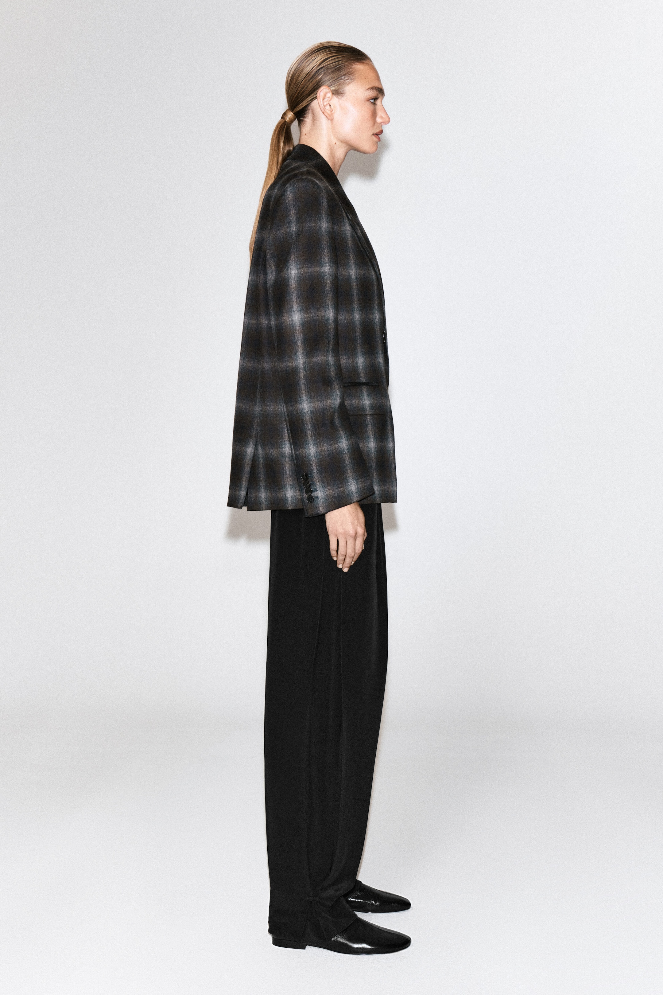 Un blazer croisé en mélange de laine à carreaux, orné d’un motif à carreaux gris foncé et brun, est porté par le mannequin, présenté de profil. Ce blazer structuré est associé à un pantalon noir à jambes larges et à des chaussures plates en cuir noir.