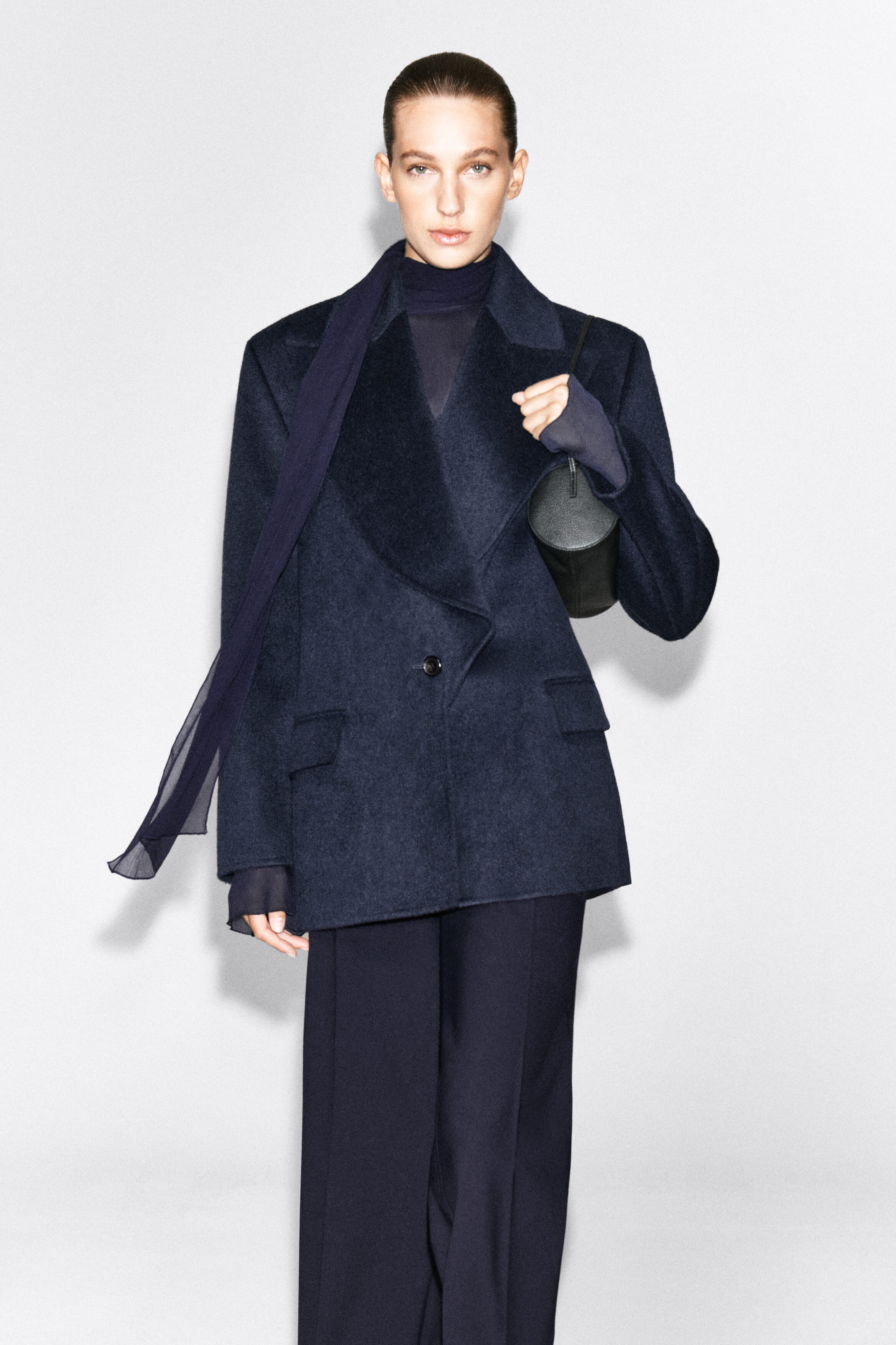 A dark navy wool-blend blazer featuring exaggerated peak lapels and front pockets is worn by the model over a sheer high-neck top and matching wide-leg trousers. The model is carrying a black cylindrical bag under one arm and wearing a long, sheer navy scarf draped over one shoulder.
