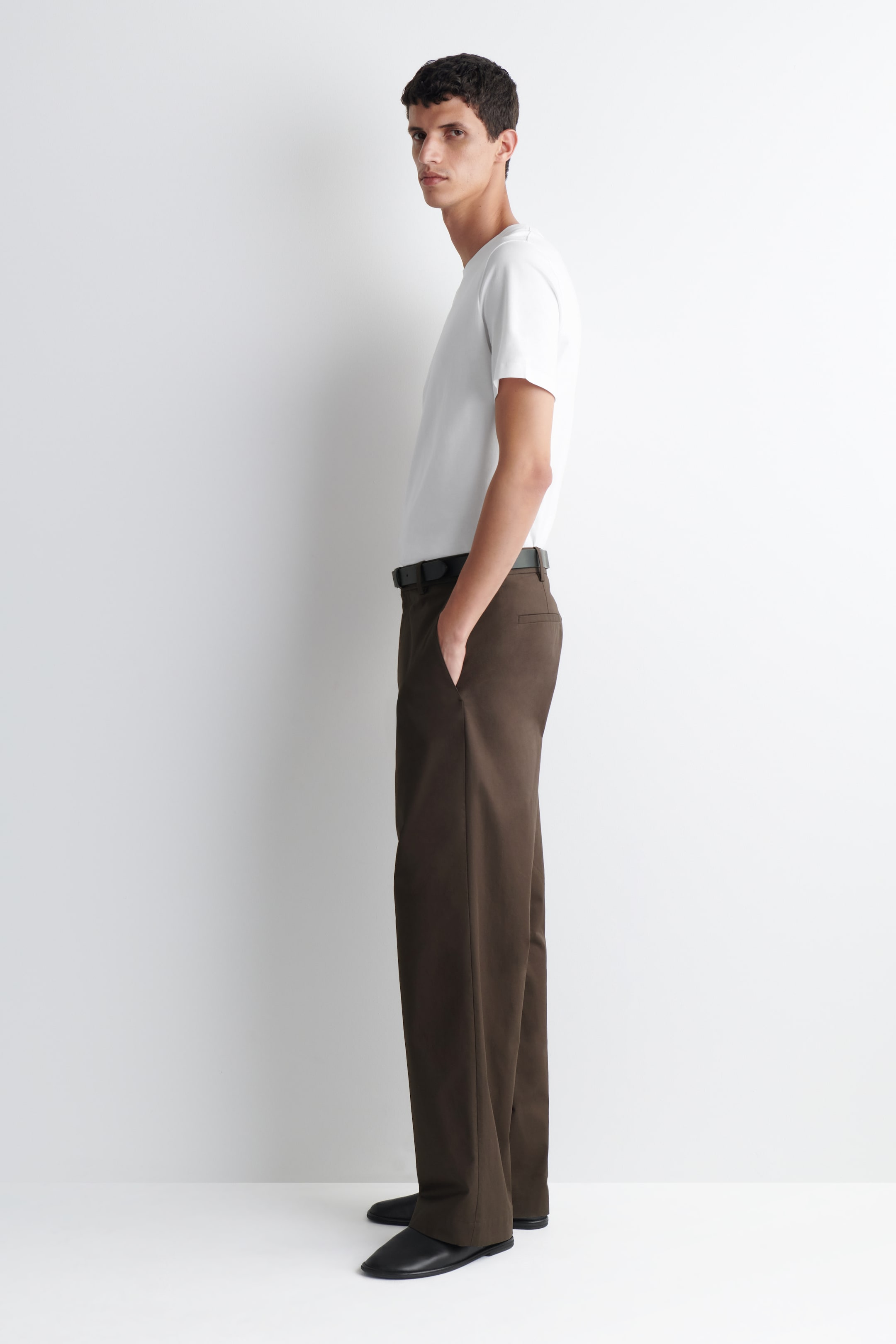 Grotere afbeelding bekijken: De bruine rechte broek wordt gedragen door het model en heeft een donkerbruine kleur. Het model staat met één hand in een voorzak, en de broek is gestyled met een zwarte riem, een wit t-shirt dat in de tailleband is gestopt, en zwarte schoenen.