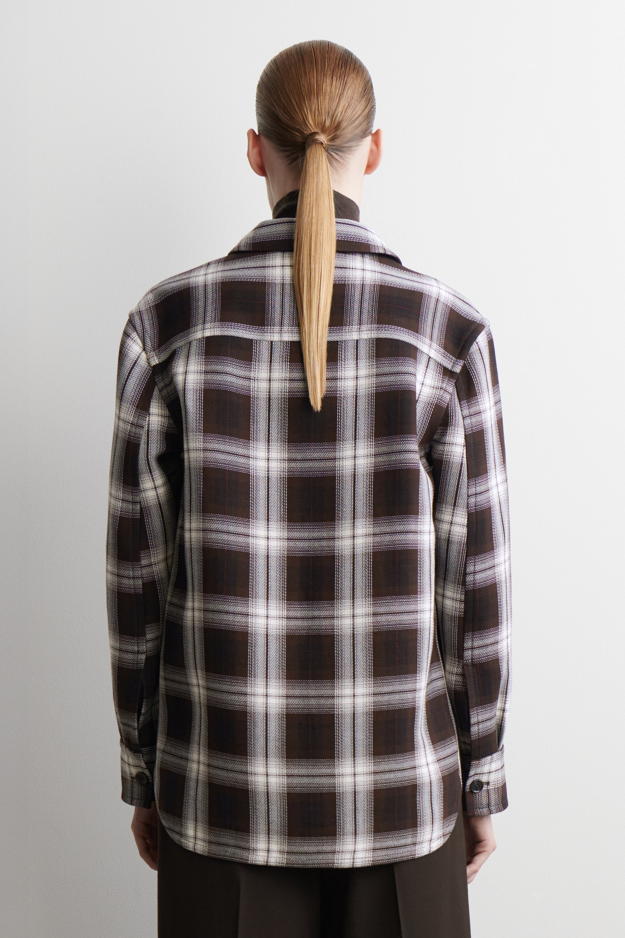 Agrandir l'image: Une chemise à carreaux marron, blanche et marron foncé est présentée de dos. Elle présente une coupe décontractée, des manches longues avec poignets boutonnés et un ourlet incurvé, et est superposée à un haut à col montant foncé visible au niveau du col.