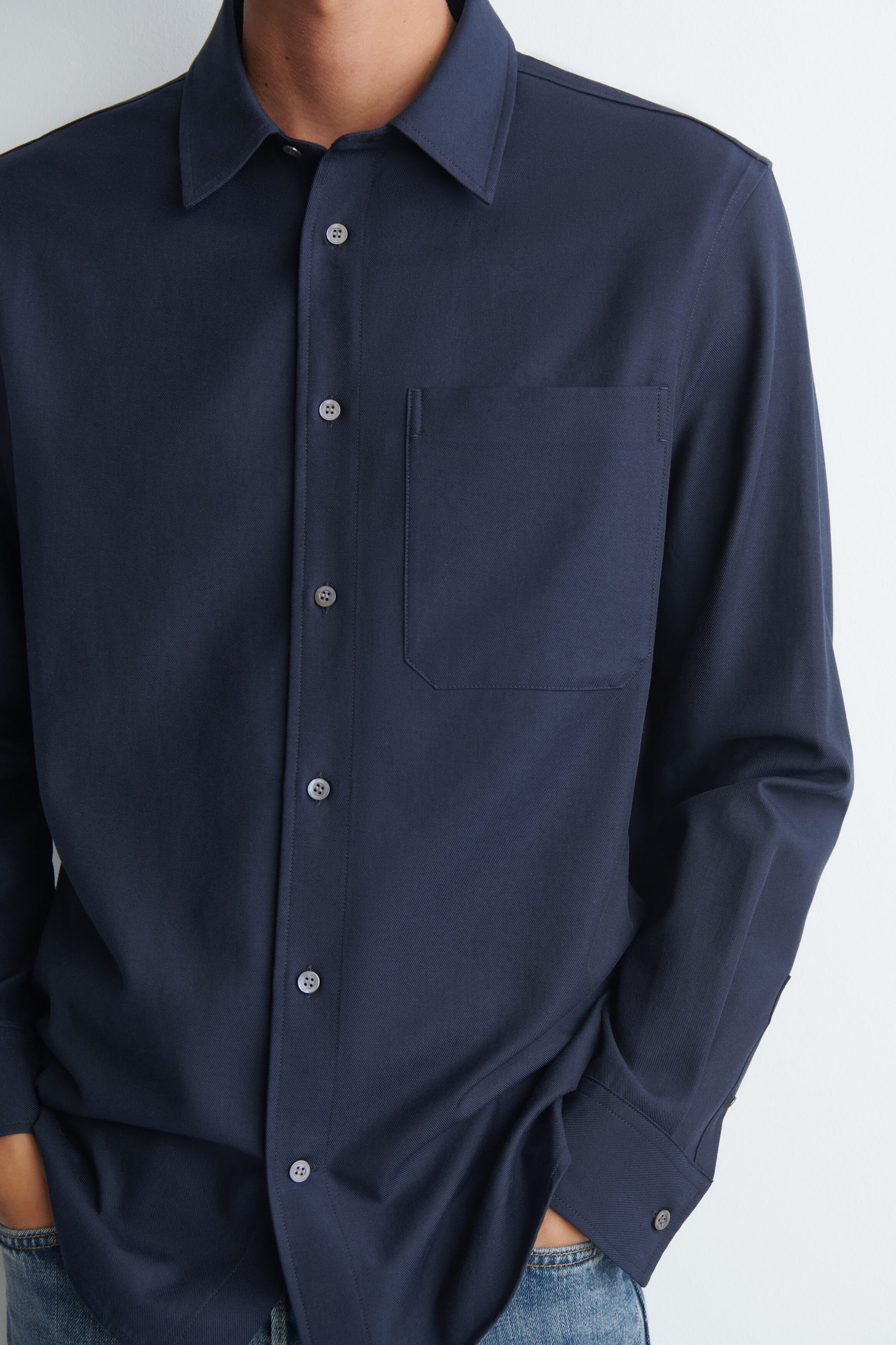 The model is wearing a navy blue relaxed twill shirt, featuring a classic pointed collar, a button-down front placket, and a single chest pocket. The shirt is styled with blue denim jeans, with the model’s hands resting in their front pockets.