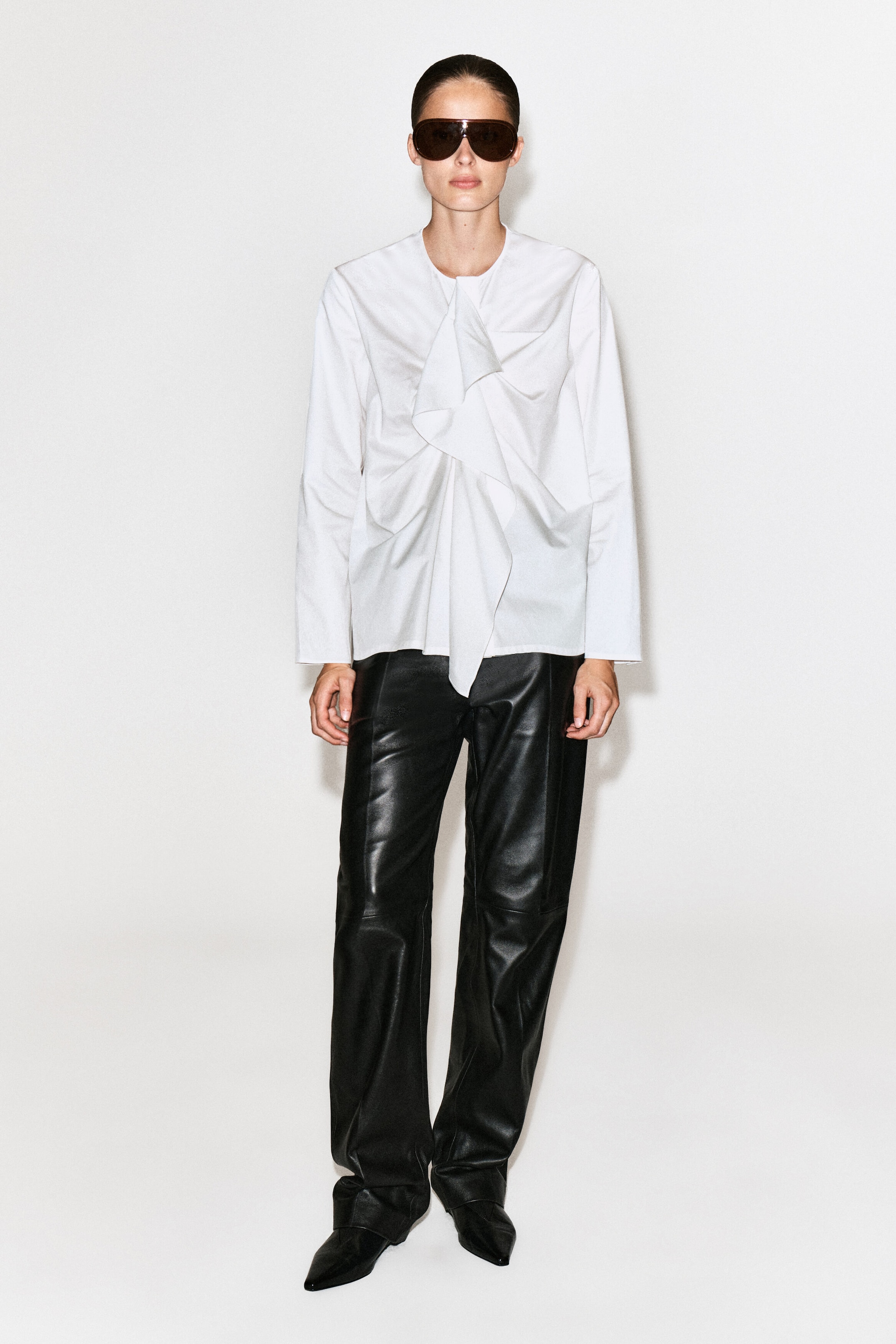 The model is wearing a white ruffled cotton blouse featuring a round neckline, long sleeves, and a prominent draped ruffle detail down the center front. The blouse is styled with black leather trousers and dark sunglasses.