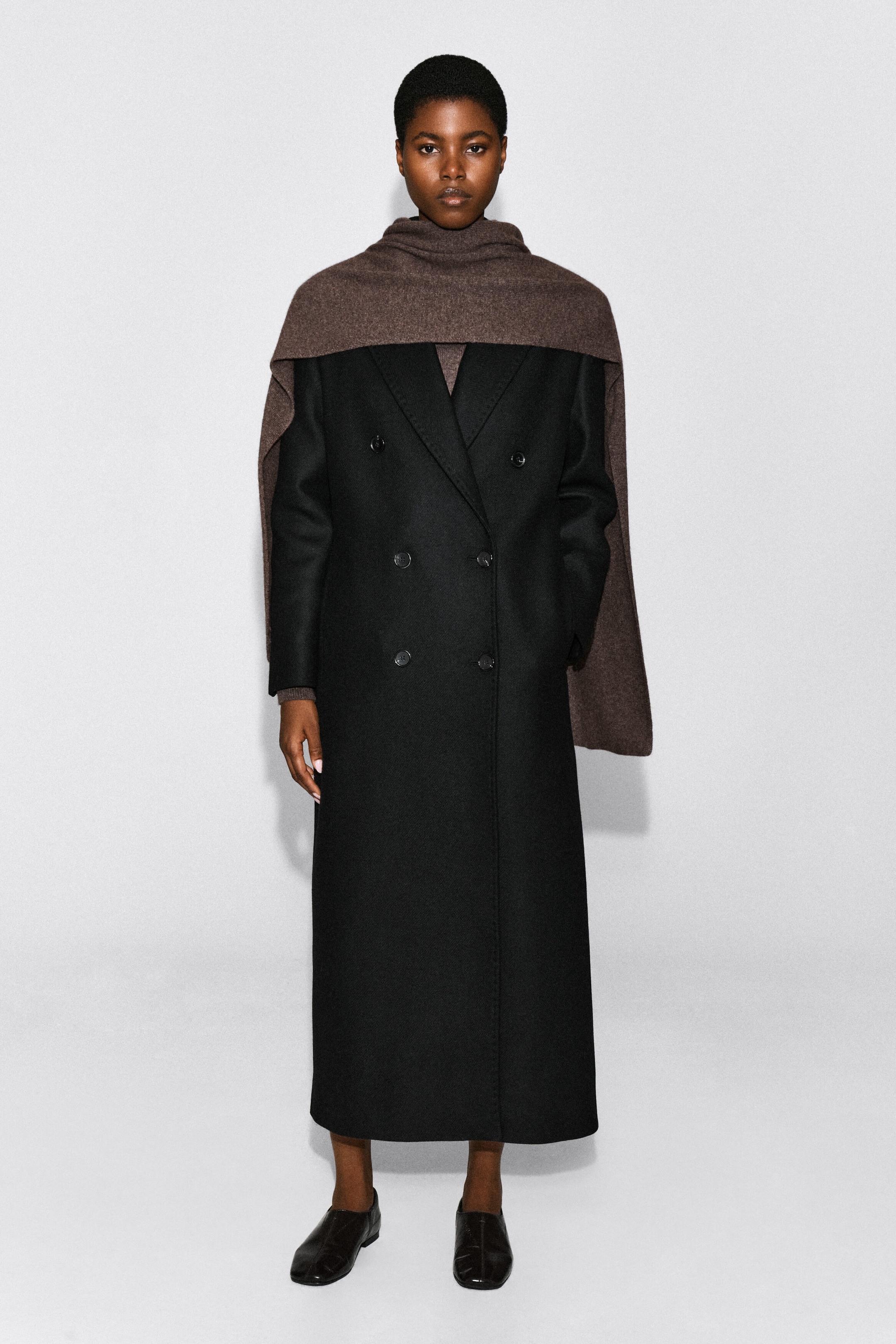 A brown cashmere scarf is worn by the model, wrapped around the neck and draped over the shoulders. The wide scarf is layered over a long black double-breasted coat. A brown cashmere scarf is worn by the model, wrapped around the neck and draped over the shoulders. The wide scarf is layered over a long black double-breasted coat.