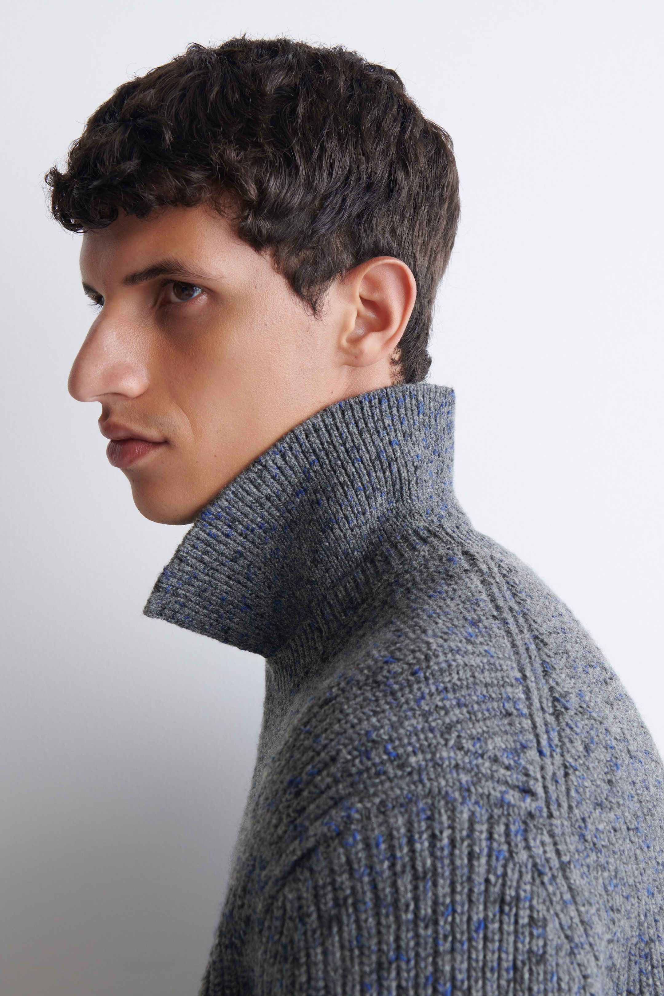 The model is wearing a grey ribbed knit turtleneck sweater with blue speckled details. The sweater features a high, structured collar and a textured knit design visible on the shoulder. The model is wearing a grey ribbed knit turtleneck sweater with blue speckled details. The sweater features a high, structured collar and a textured knit design visible on the shoulder.