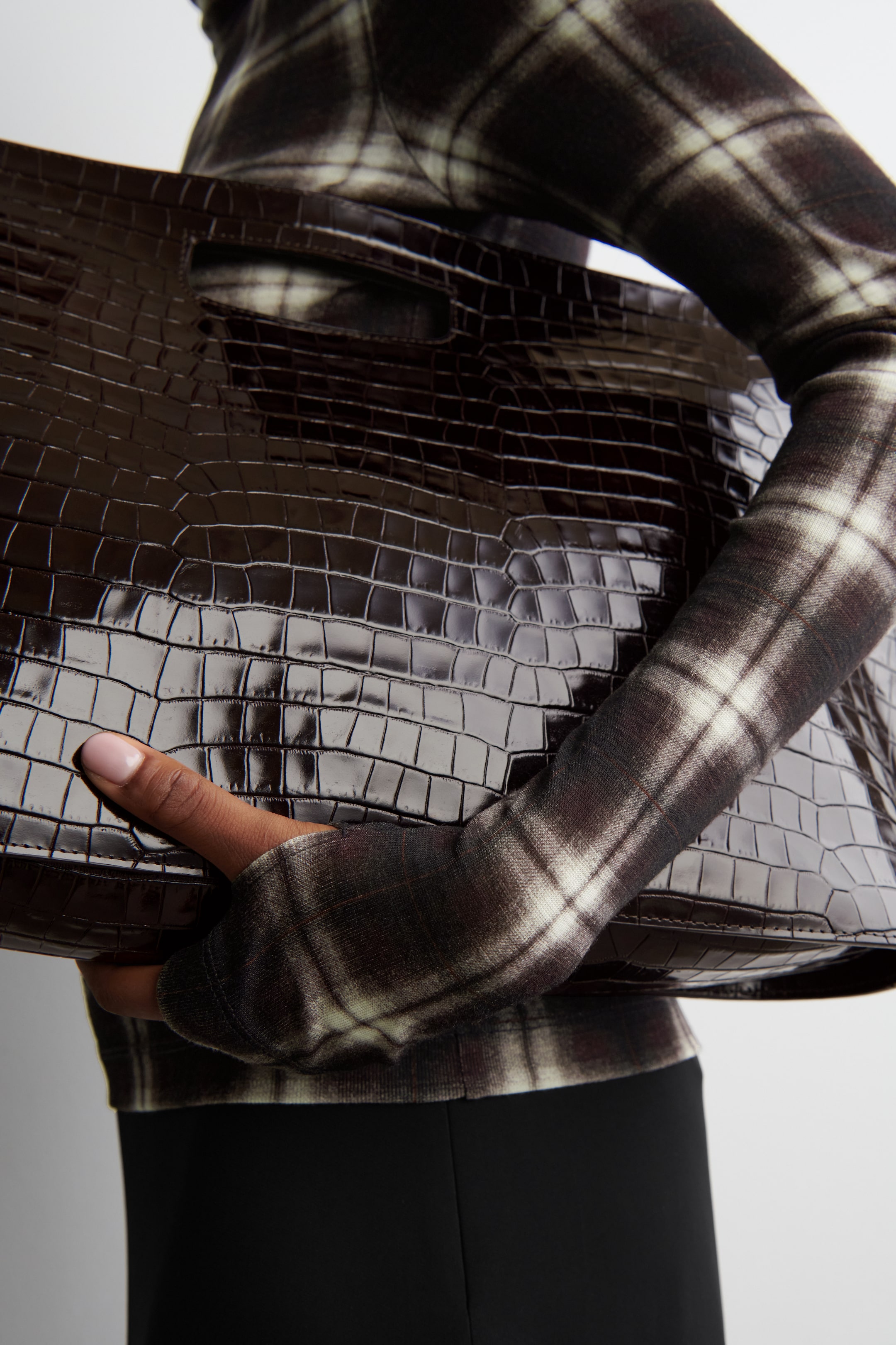 Agrandir l'image: Un sac fourre-tout en cuir embossé crocodile brun foncé et brillant avec des poignées découpées est tenu par une personne, avec ses surfaces avant et supérieure visibles. Le sac complète un haut à manches longues avec un motif à carreaux foncés et clairs.