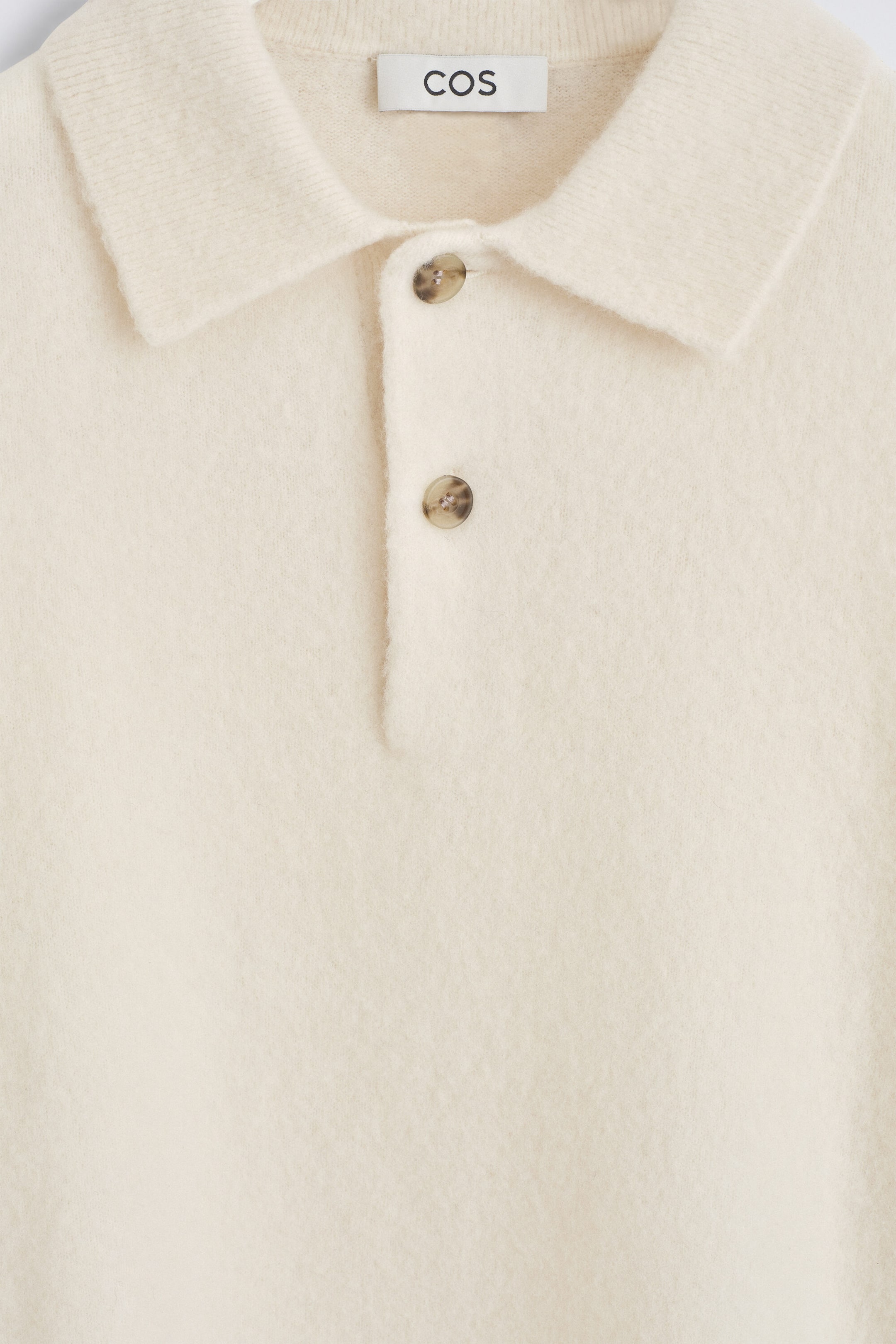 This close-up front view of a cream-colored brushed wool polo shirt features a classic knit collar and a partial placket with two tortoiseshell-patterned buttons. The soft, textured fabric shows a COS brand label sewn into the inner neckline.