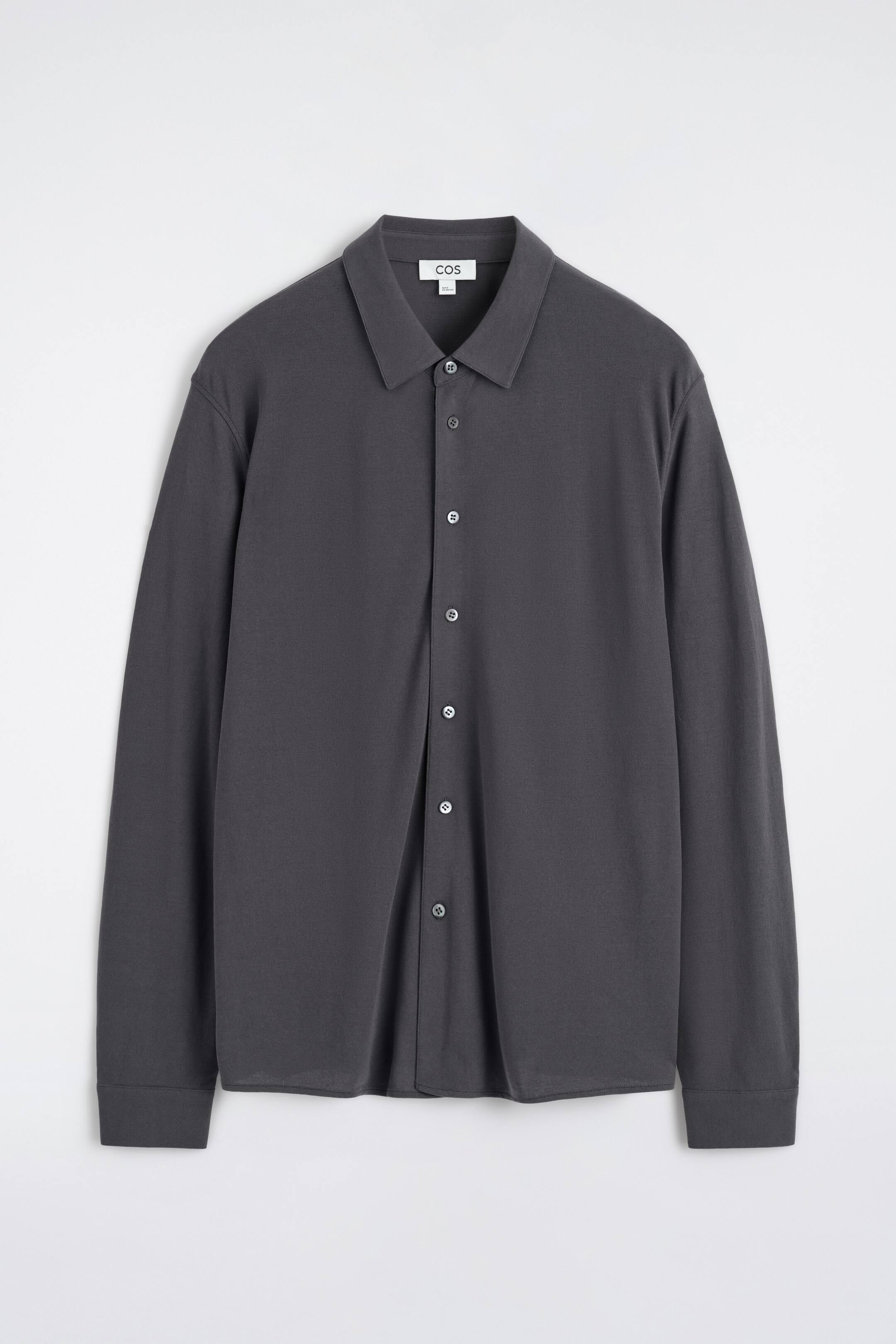 Zobrazit větší obrázek: Představujeme tmavě šedou košili s dlouhým rukávem, která je zobrazena zepředu. Má špičatý límec, plnou knoflíkovou légu se světlými knoflíky a manžety na jeden knoflík.