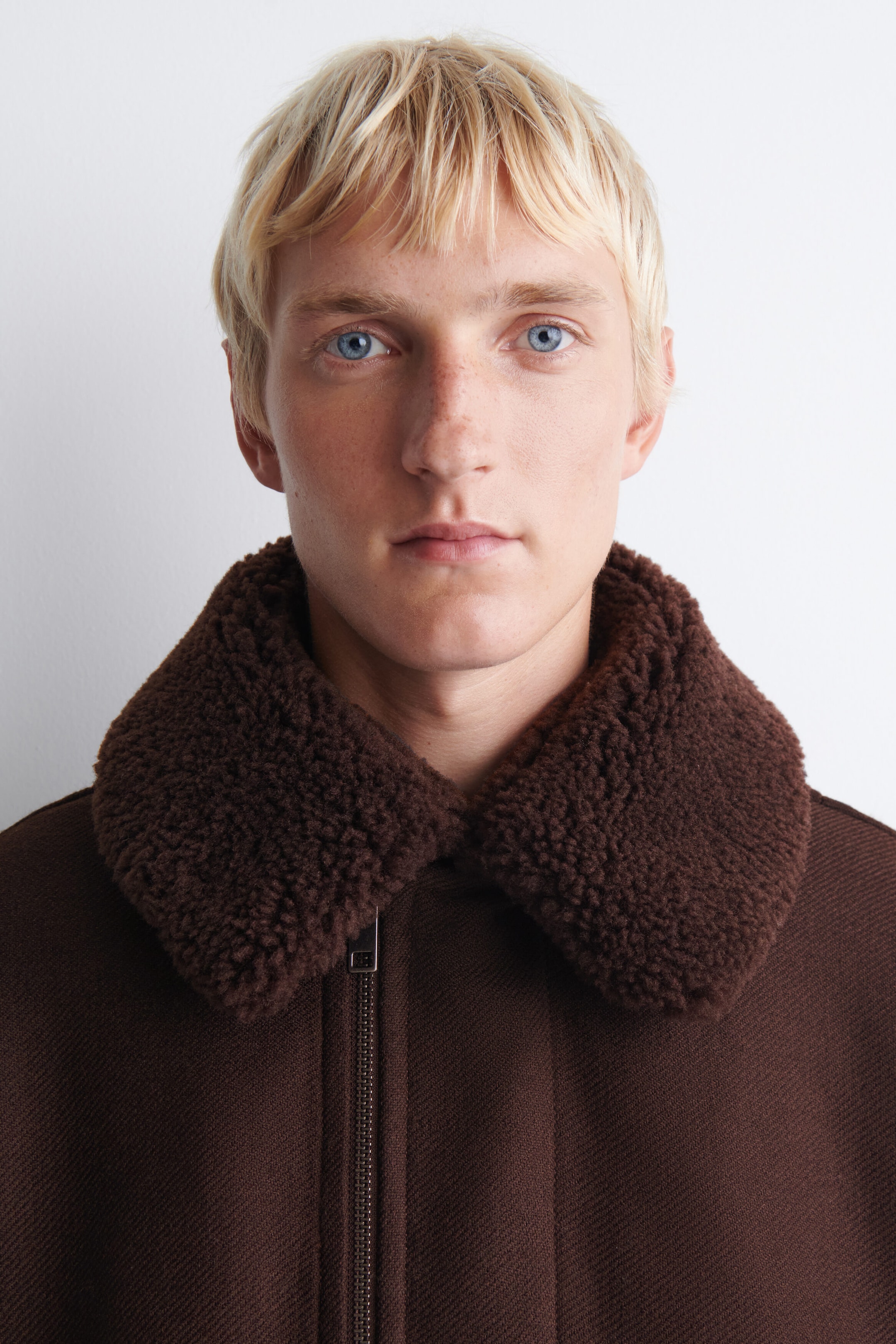 A dark brown jacket features a plush, textured shearling collar and a metallic front zipper on a textured woven fabric, shown from the chest up. A dark brown jacket features a plush, textured shearling collar and a metallic front zipper on a textured woven fabric, shown from the chest up.