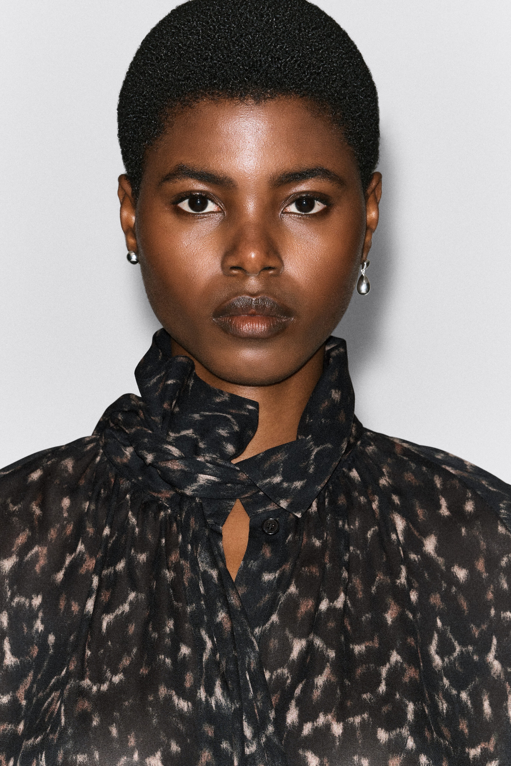A leopard-print fluid top featuring a high collar with an integrated, knotted scarf detail is being worn by the model. The lightweight fabric has a dark abstract leopard pattern and a small button closure visible at the neckline. A leopard-print fluid top featuring a high collar with an integrated, knotted scarf detail is being worn by the model. The lightweight fabric has a dark abstract leopard pattern and a small button closure visible at the neckline.