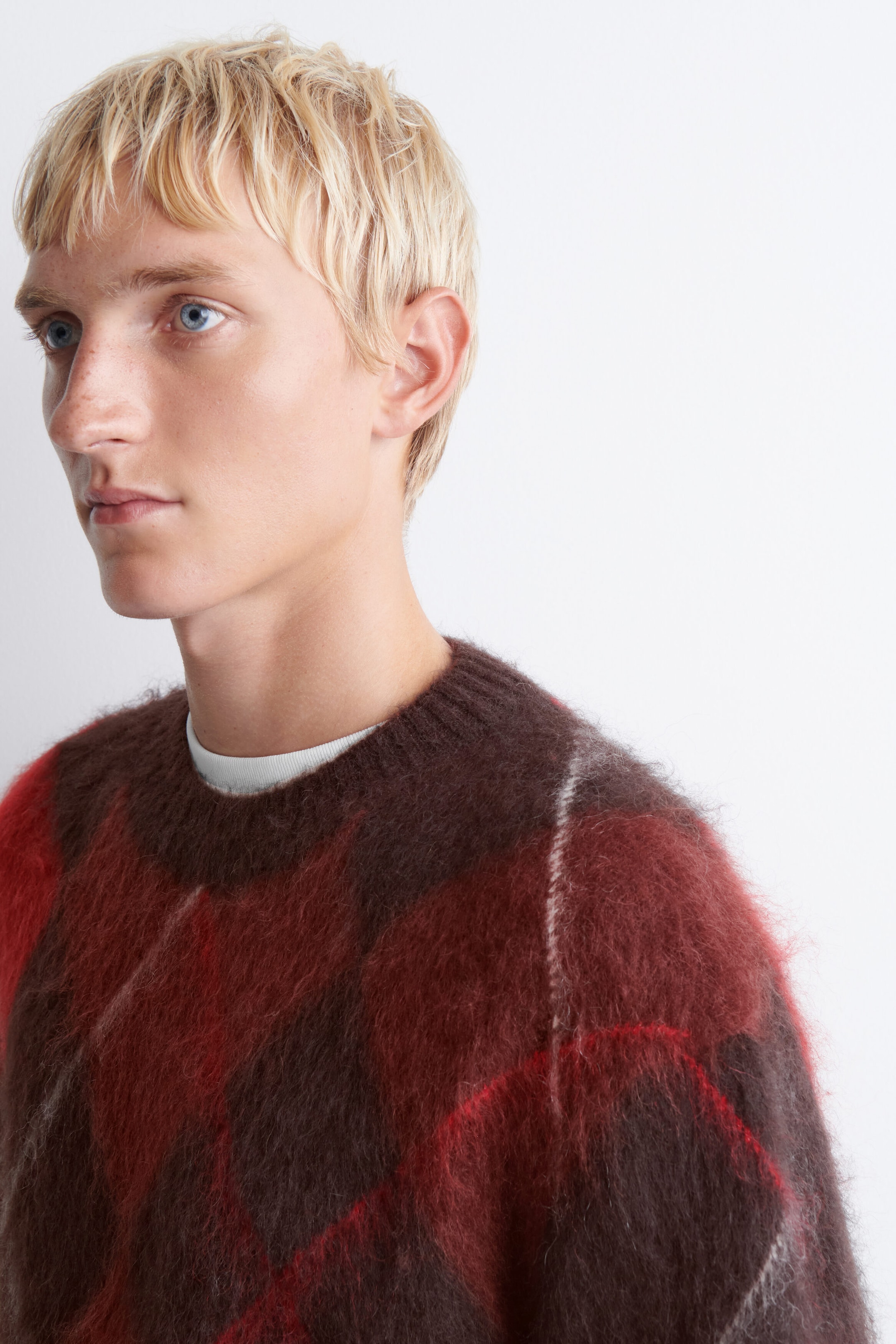 Dieser flauschige, aufgerauhte Strickpullover im Argyle-Muster zeigt ein braunes und rotes Rautenmuster mit geripptem Rundhalsausschnitt. Er wird in Lagen über einem weissen T-Shirt mit Rundhalsausschnitt getragen, das am Ausschnitt sichtbar ist.