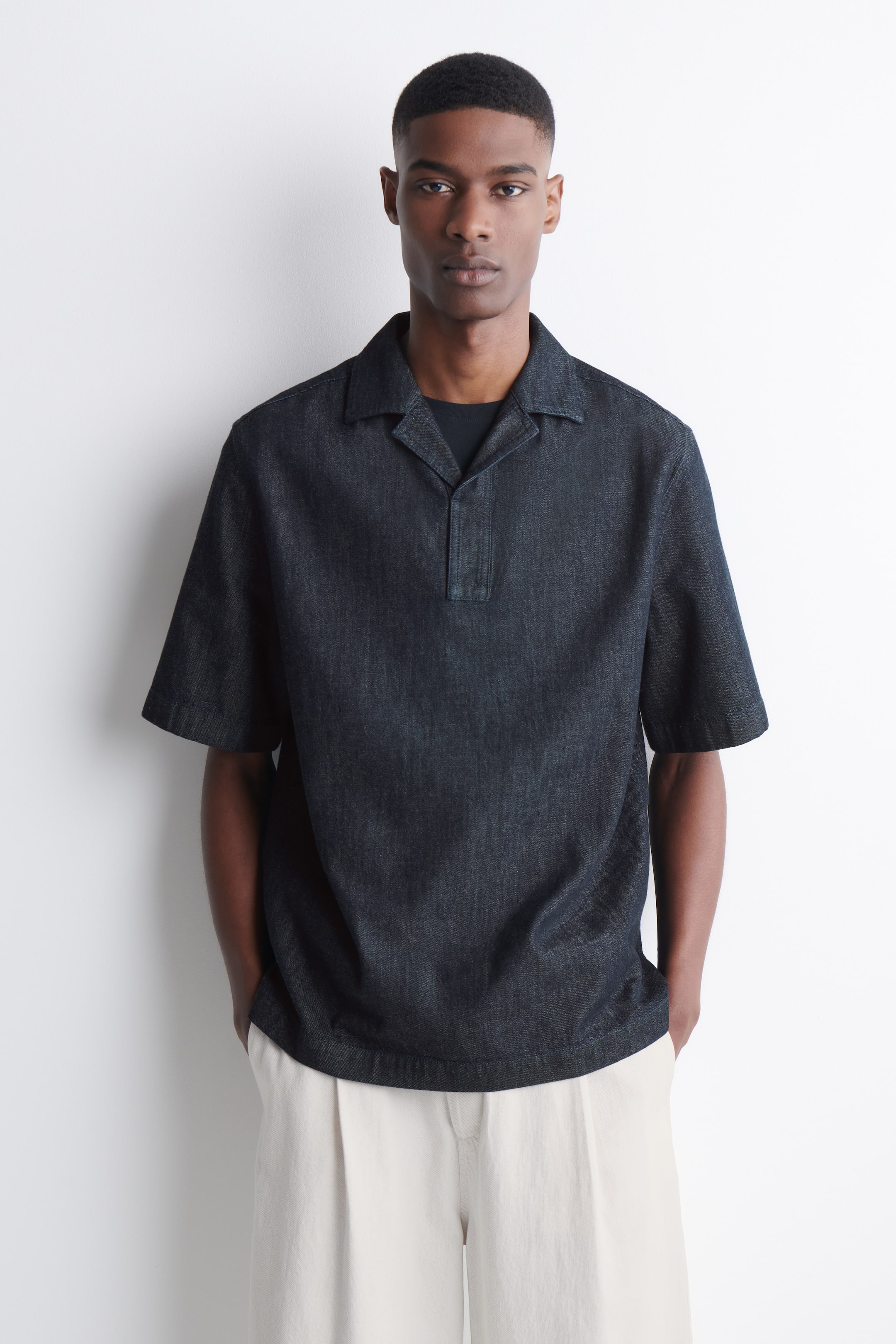 הצגת תמונה גדולה יותר: חולצה כהה עם שרוולים קצרים עשויה דנים, מציגה צווארון קמפ ופליקט חצי תפור. היא מציעה גזרה רגועה עם שרוולים קצרים ורחבים, בעוד חולצה שחורה נראית מתחת לצווארון הפתוח שלה.