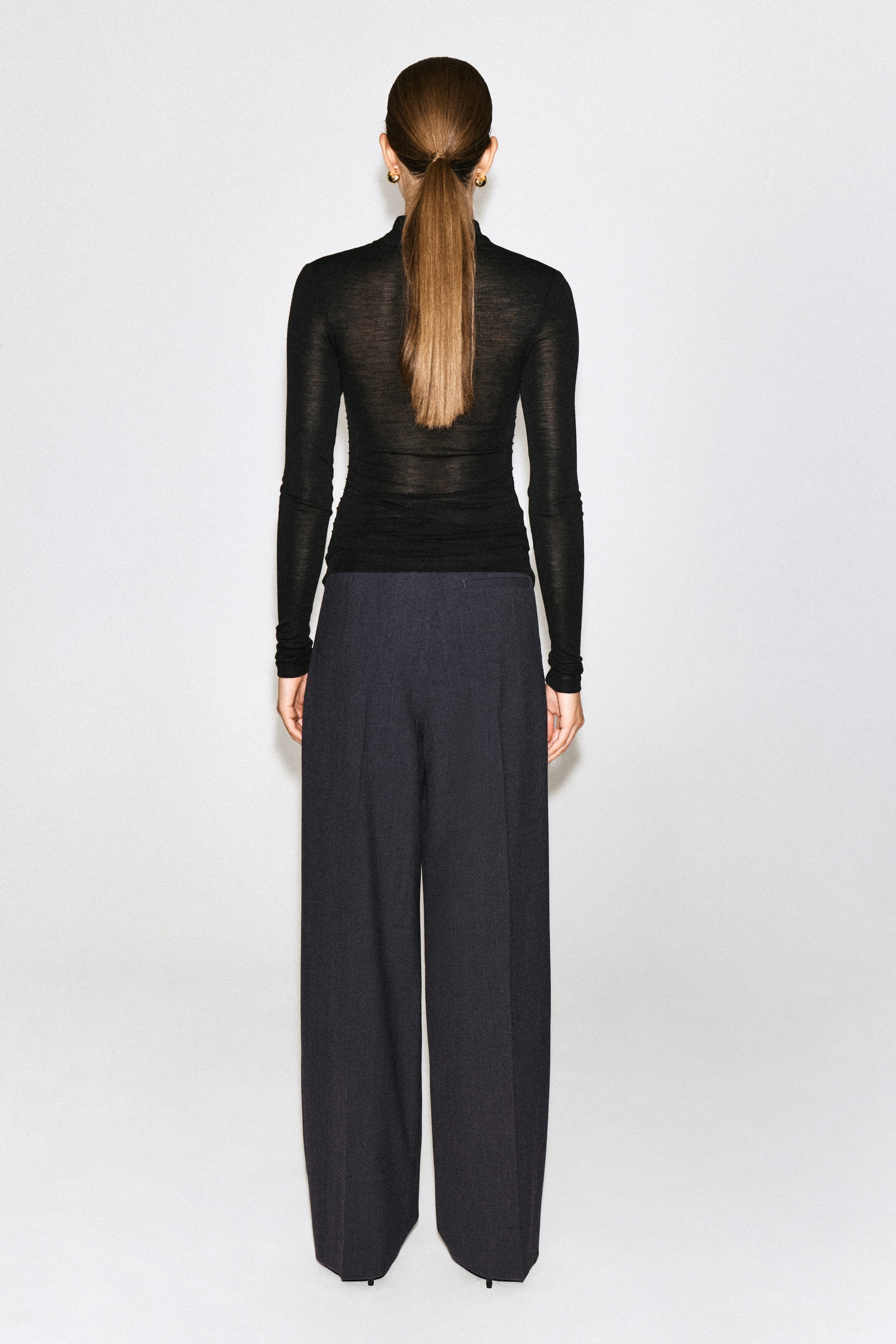 A black, semi-sheer, long-sleeved knit top is being worn by the model, who is facing away from the camera. The slim-fit top features a high collar and is paired with dark, wide-leg trousers. A black, semi-sheer, long-sleeved knit top is being worn by the model, who is facing away from the camera. The slim-fit top features a high collar and is paired with dark, wide-leg trousers.