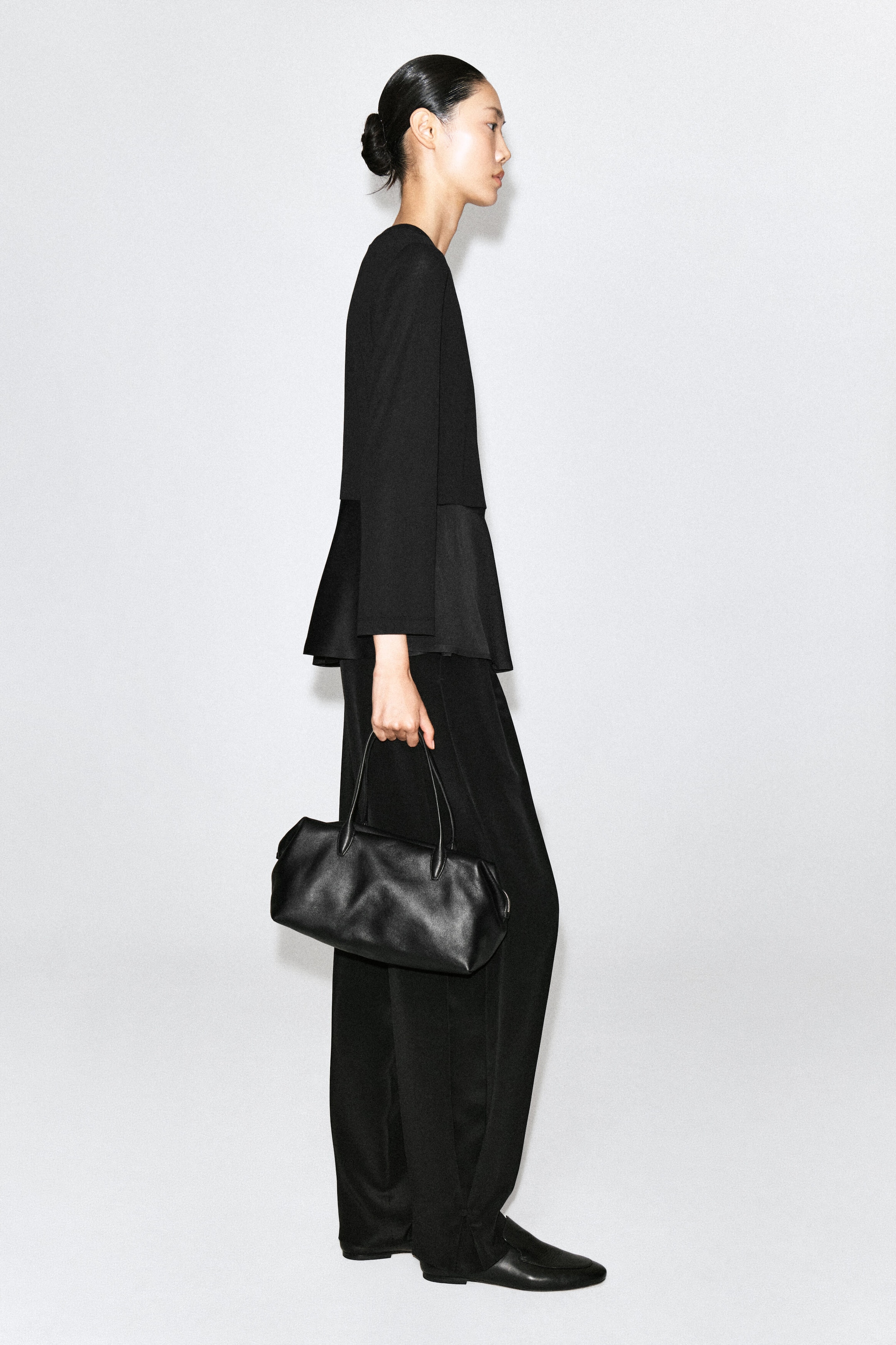 Un haut en jersey peplum satiné noir, doté de manches longues et d’un ourlet évasé en satin contrastant, est porté par le mannequin. Le haut est agencé avec un pantalon à jambe large, tandis que le mannequin tient un sac à main en cuir noir.