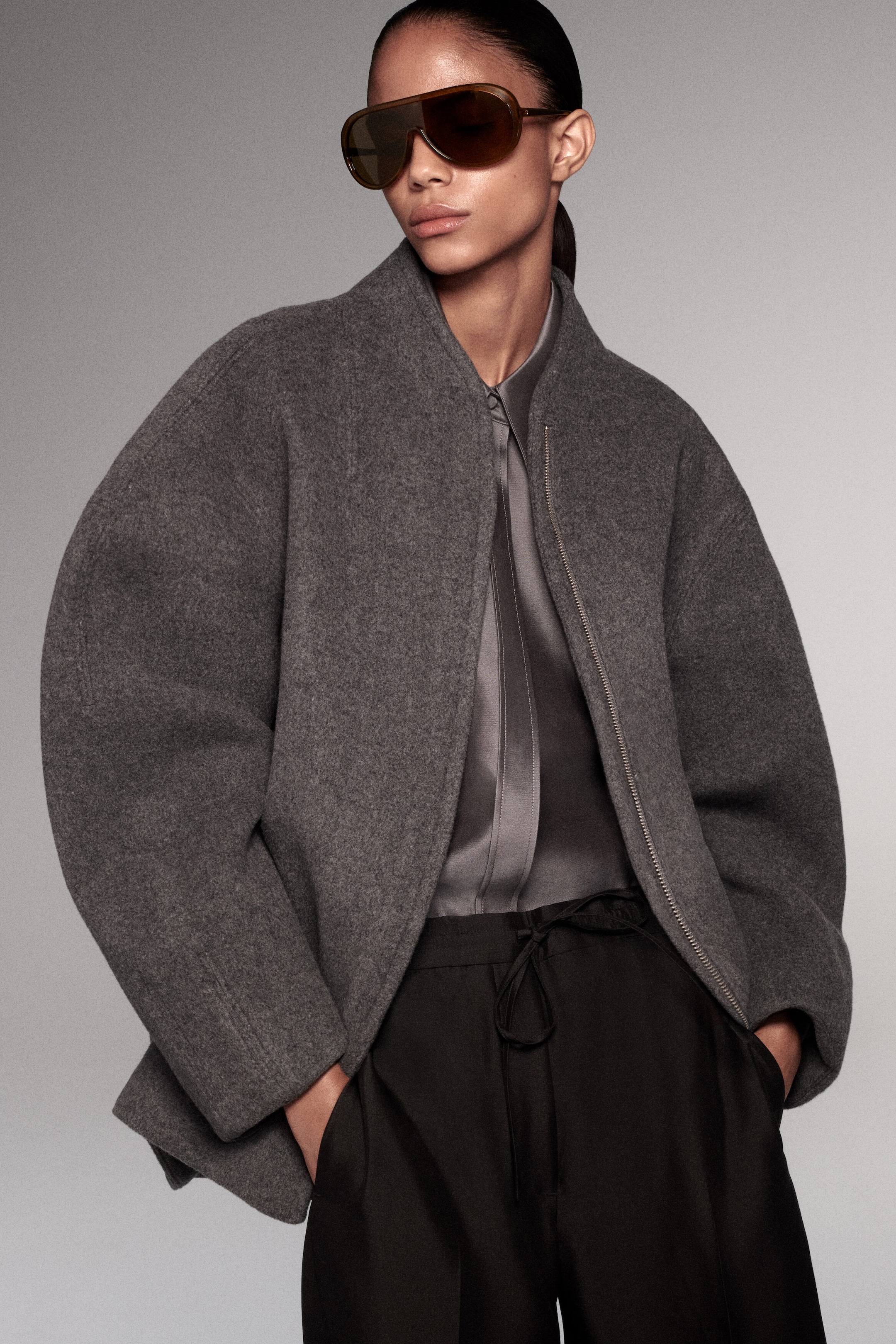 Cette veste zippée en mélange de laine sculpté, en gris texturé, est présentée de face, mettant en valeur une silhouette surdimensionnée et arrondie, avec des manches cocon volumineuses et un col montant. La veste est portée partiellement ouverte sur une chemise en satin gris et un pantalon noir, soulignant son tombé structuré et lourd.