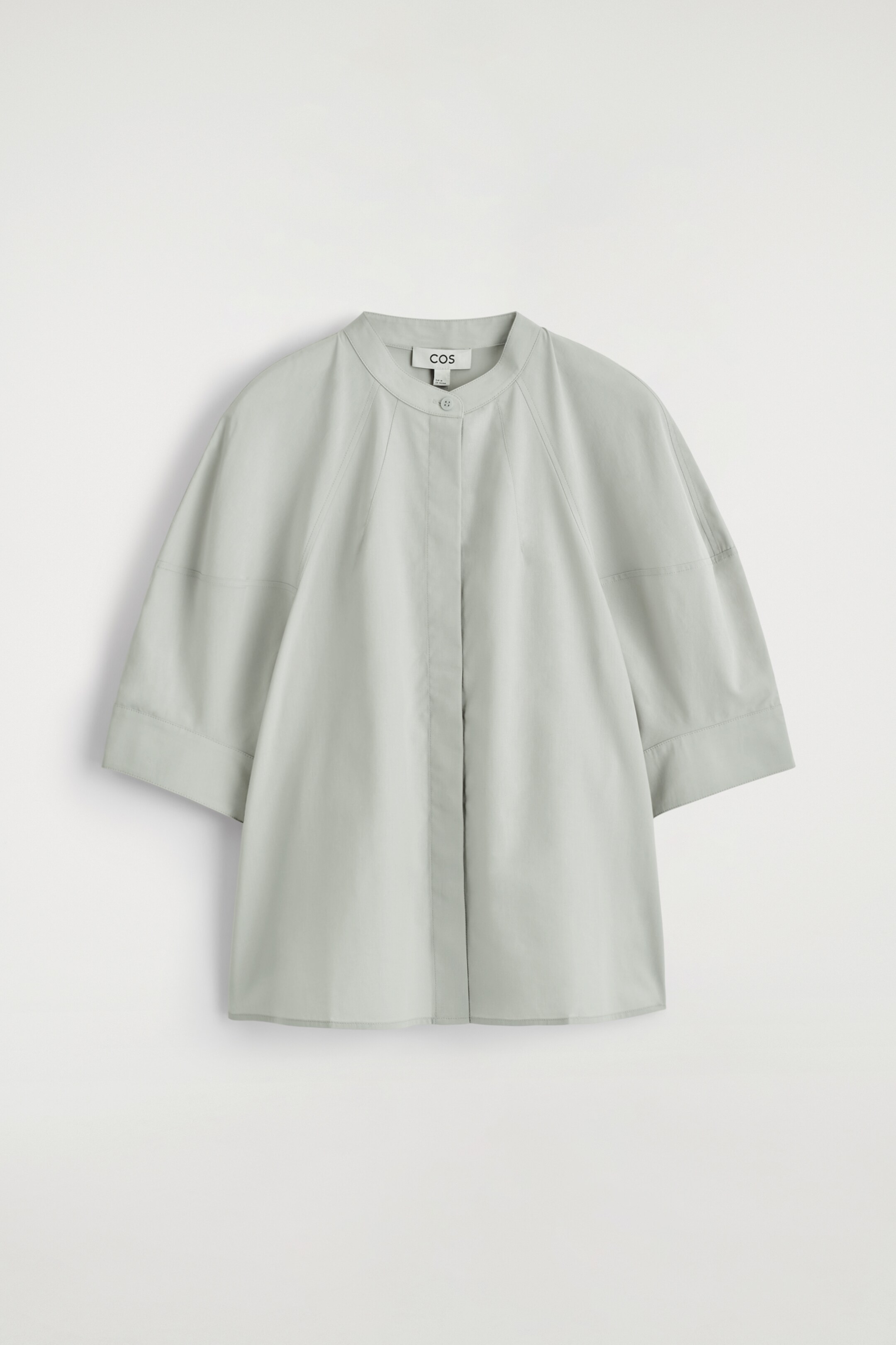 Grotere afbeelding bekijken: Een lichtgroene blouse ligt plat, gepresenteerd van de VOORKANT. Het heeft een ronde grandad-kraag, een verborgen knoopsluiting en volumineuze raglanmouwen die boven de elleboog eindigen. De blouse loopt subtiel uit naar de zoom.