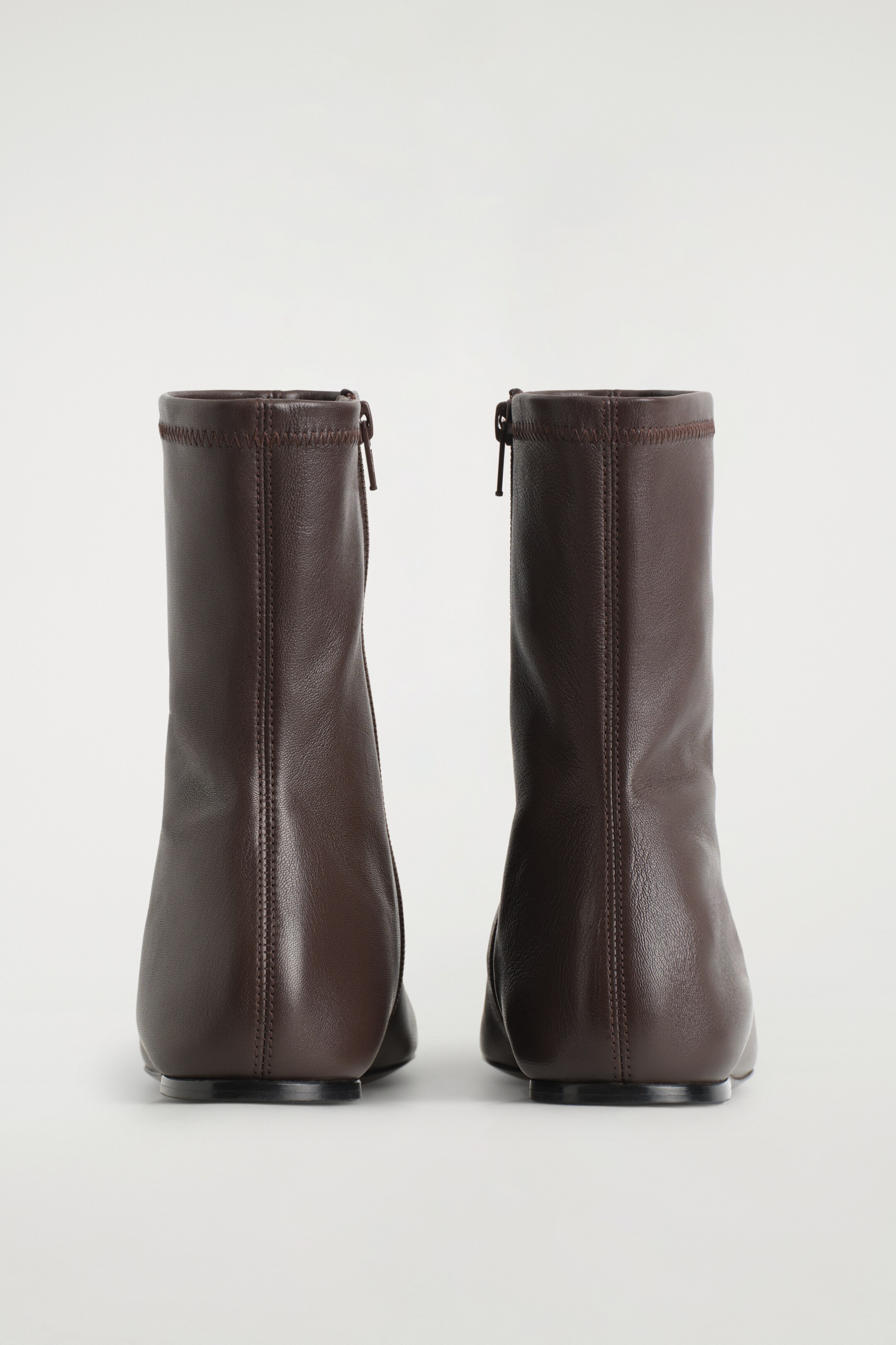 View larger image: A pair of dark brown leather sock boots is facing BACK, each featuring a vertical metal zipper with an elongated pull tab on the shaft. Elasticized stitching is detailing the top edge, a central vertical seam is running down the back, and the boots are presenting a sleek silhouette with flat heels.
