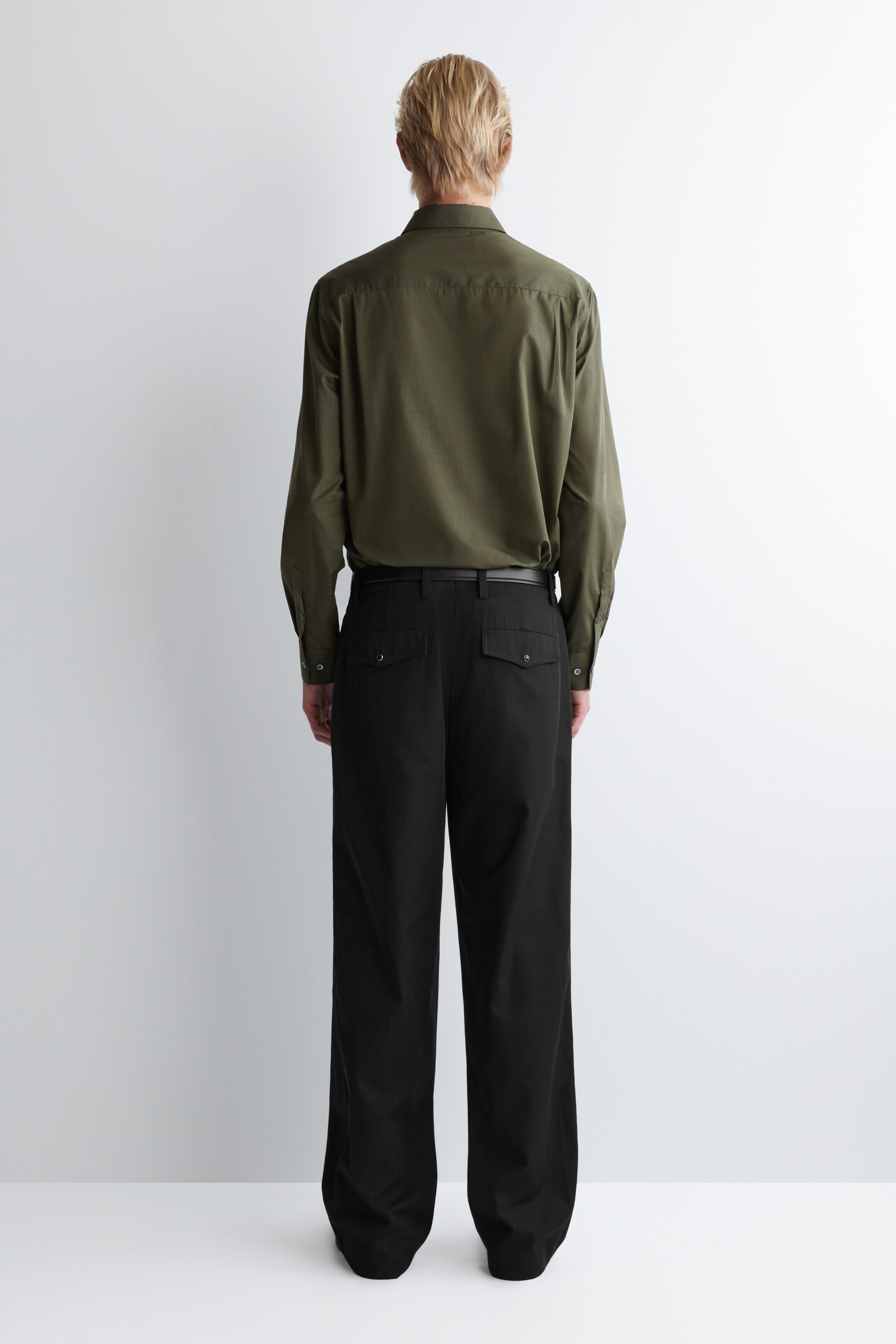 Visualizza immagine più grande: Una camicia a maniche lunghe verde oliva, caratterizzata da un carrello posteriore e polsini con bottoni, è indossata, presentata di dietro, ed è infilata nei pantaloni neri a gamba larga. I pantaloni neri presentano una cintura con una cintura nera, passanti per la cintura e due tasche posteriori con patta e bottone.