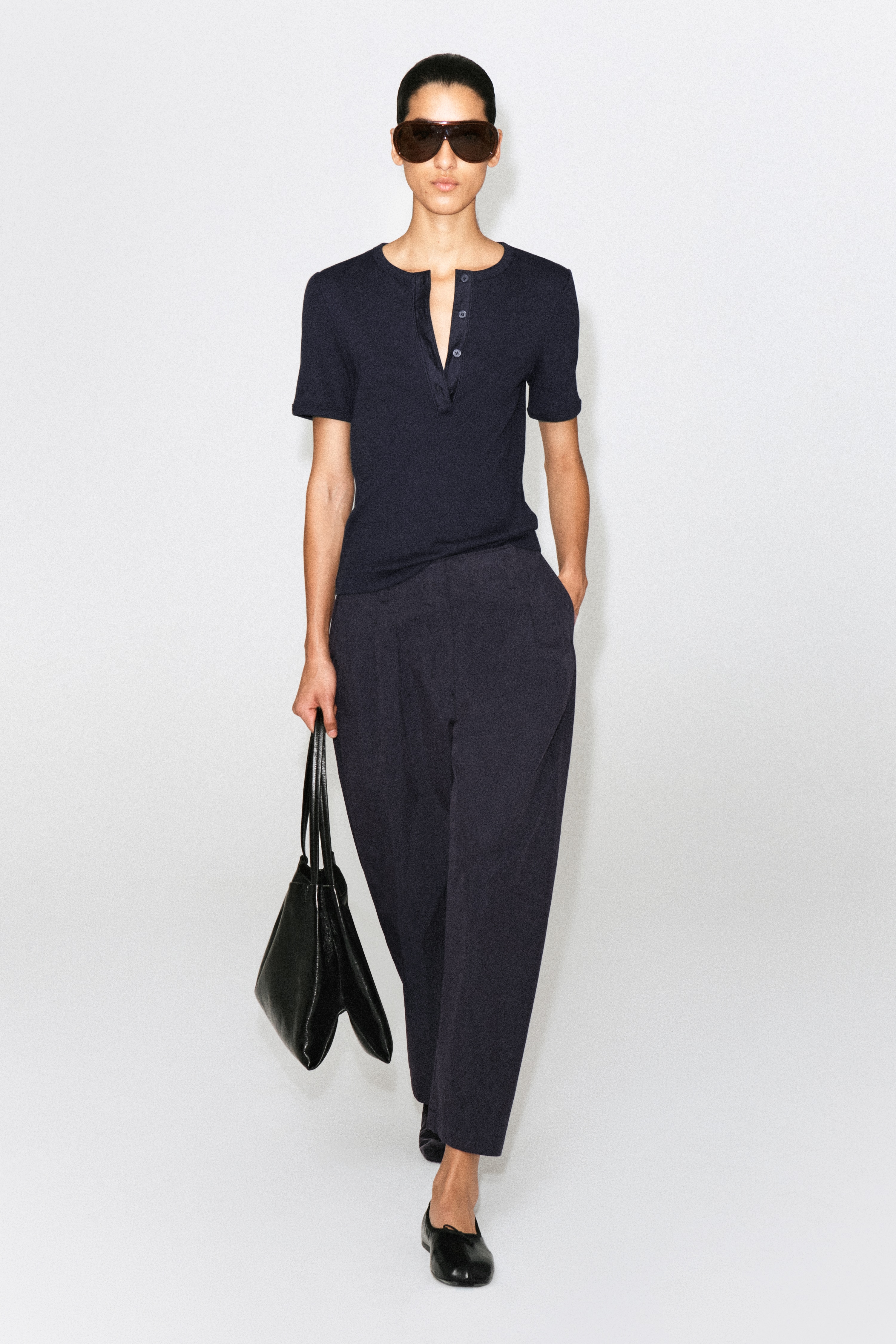 Donkerblauwe cropped broek wordt van voren getoond, met een hoge taille en riemlussen. Overdreven plooien zijn zichtbaar aan de voorkant, waardoor een brede, ballonvormige pijp ontstaat.