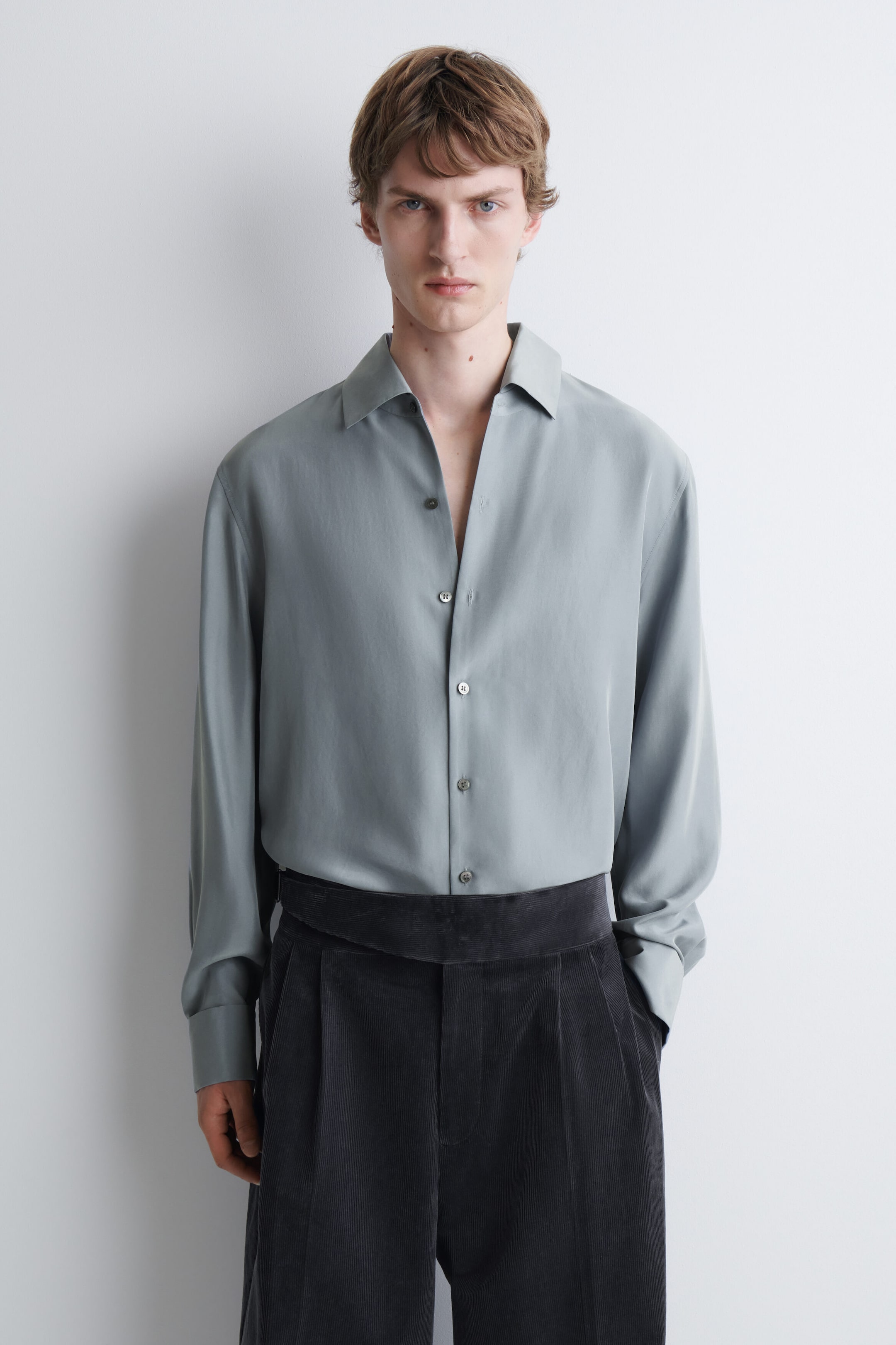 A relaxed silk shirt in light sage green is worn by the model, featuring a pointed collar, long sleeves, and a button-down front with the top buttons left open. The shirt is tucked into a pair of high-waisted, dark grey corduroy trousers. A relaxed silk shirt in light sage green is worn by the model, featuring a pointed collar, long sleeves, and a button-down front with the top buttons left open. The shirt is tucked into a pair of high-waisted, dark grey corduroy trousers.