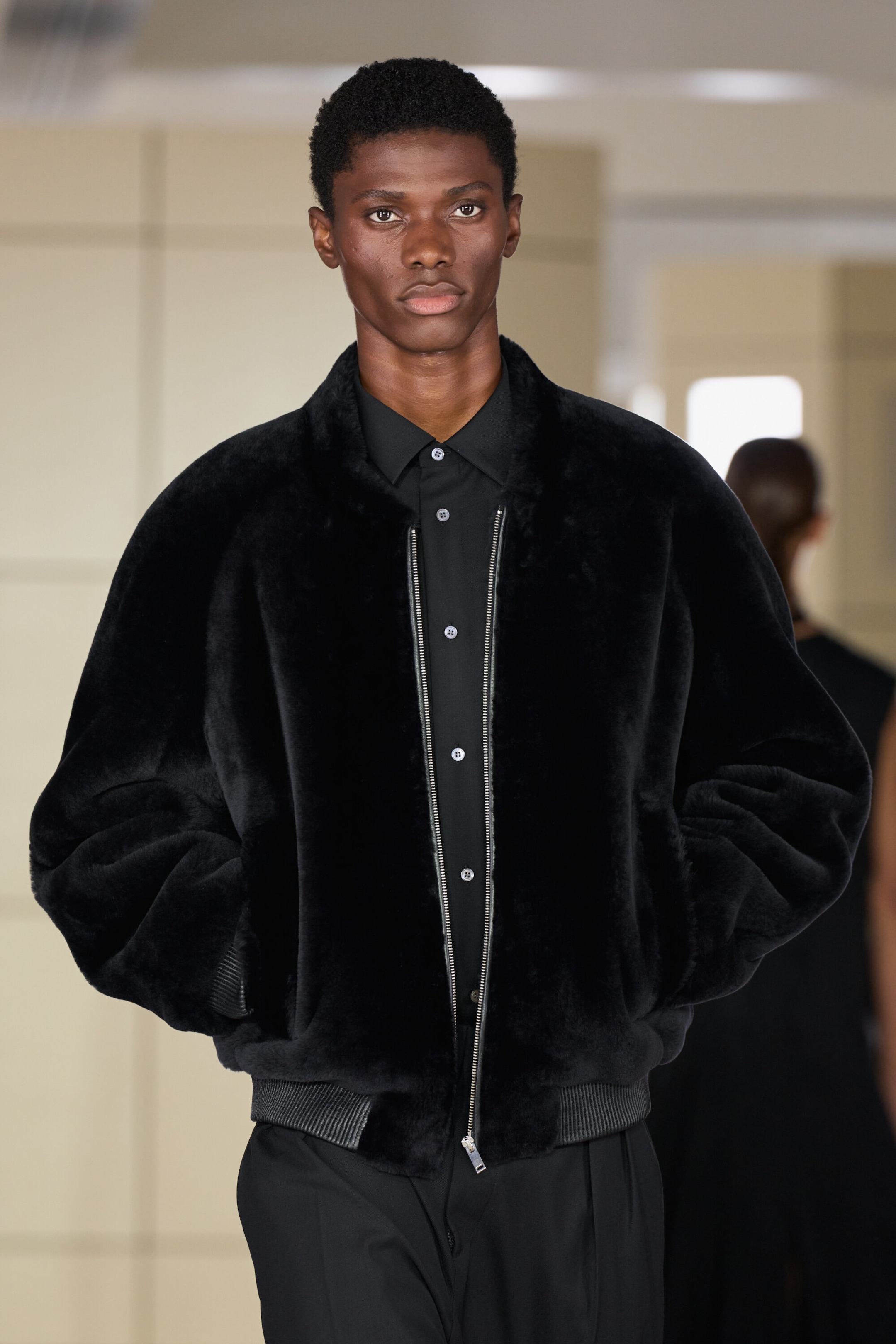 The padded shearling bomber jacket is shown from the front, featuring a plush black shearling exterior with a voluminous, relaxed fit. It is styled partially unzipped over a black button-up shirt, highlighting its ribbed hem and silver front zipper.