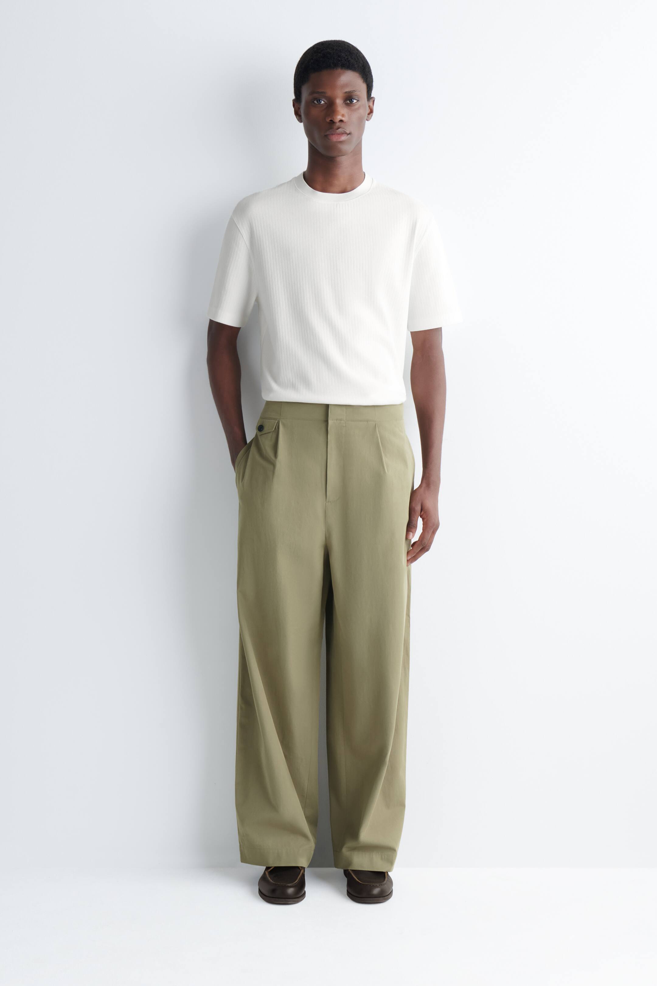 Grotere afbeelding bekijken: Een crèmekleurig geribd t-shirt met korte mouwen wordt gedragen met olijfgroene broek met wijde pijpen. De broek heeft voorplooien, een hoge taille en een zichtbare knoopsluiting aan de taille, en valt losjes over bruine schoenen.